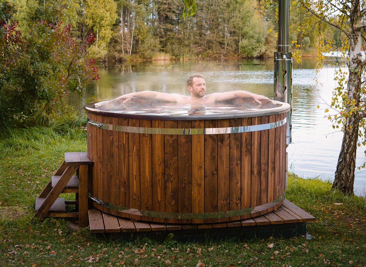 A person relaxing in a wood-fired Makoya Spa hot tub SOPHIA by a lakeside, featuring a metal chimney, wooden entry steps, and a forest in the backround.