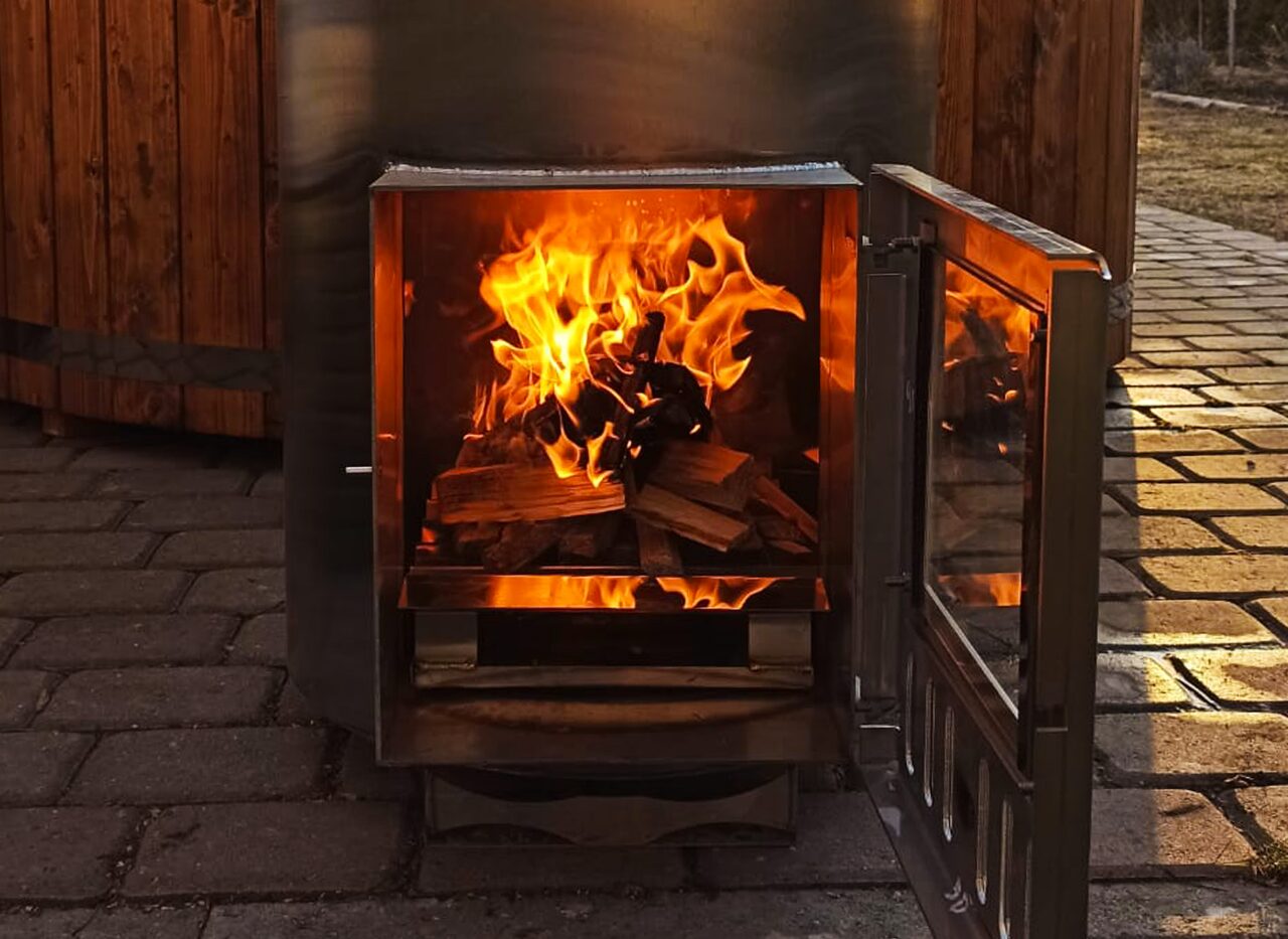 Dark metal stove attached to a Makoya Spa hot tub with the door open. Burning wood is visible.