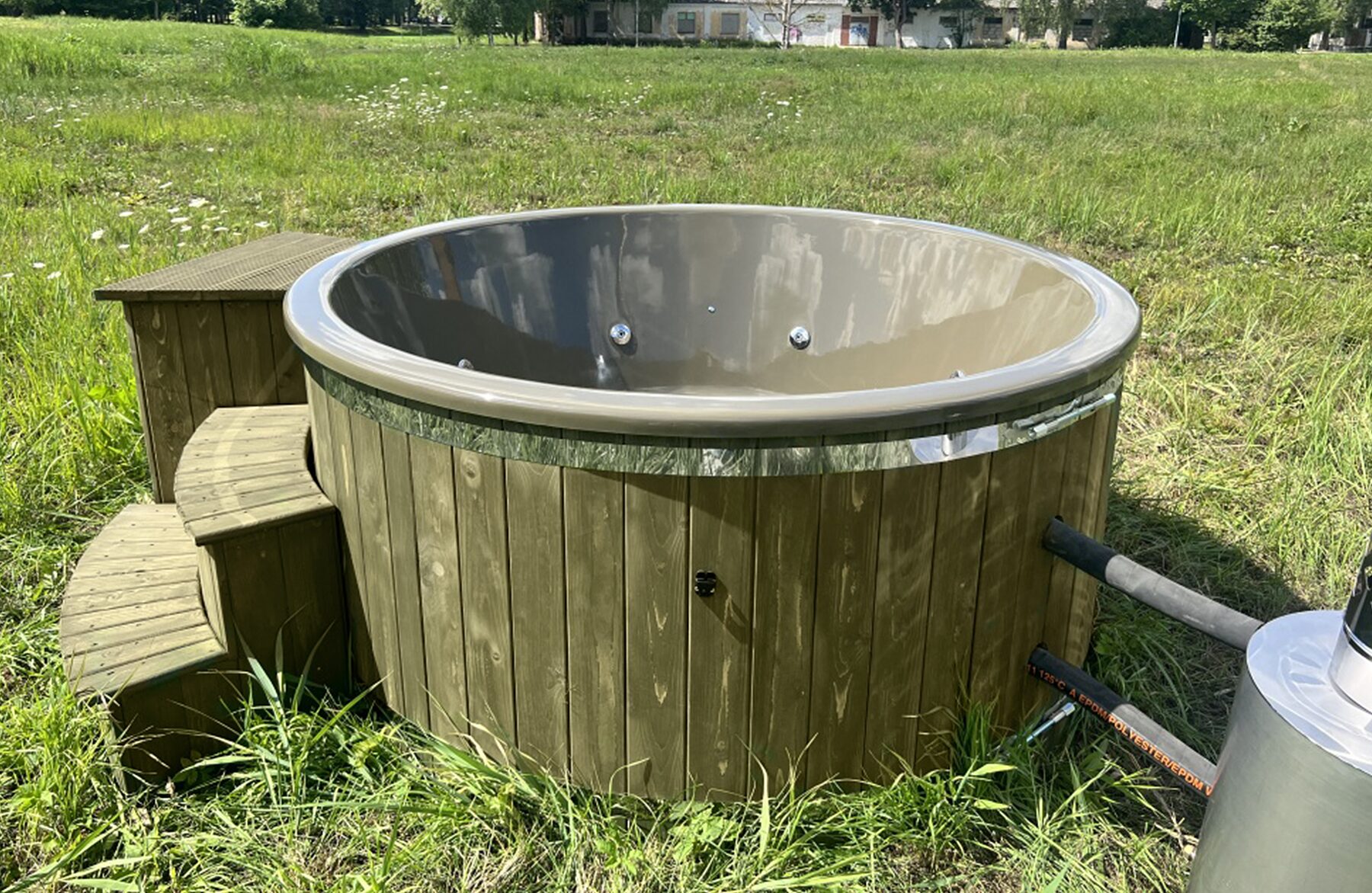 Makoya Spa hot tub DIANA set on a meadow featuring wooden cladding, a grey interior tub with integrated jets, wooden. Entry step and a external metal stove