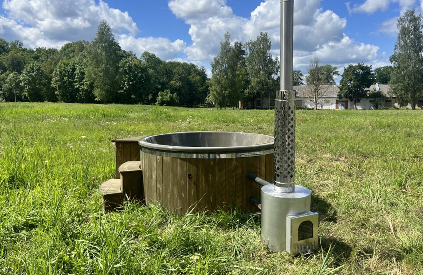 Makoya Spa Hot Tub DIANA steht auf einer grünen Wiese mit Holzverkleidung, einem externen Metallofen mit Schornstein sowie einer Holz-Einstiegstreppe, umgeben von Bäumen und bewölktem Himmel