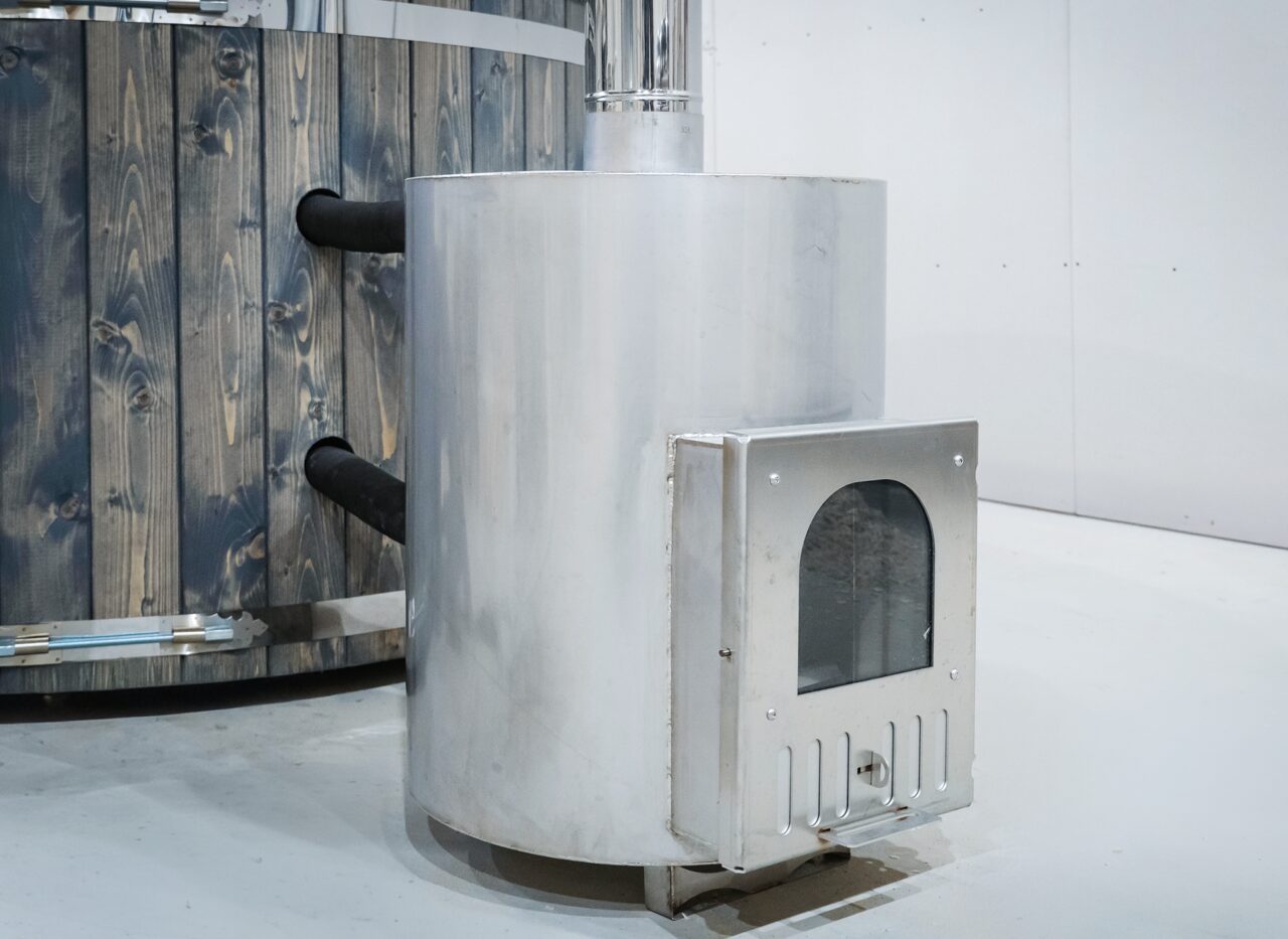 Close-up view of an external metal stove of the Makoya Spa hot tub DIANA, featuring the firebox door, connection pipes to the tub and a robust construction next to the wooden cladding. 