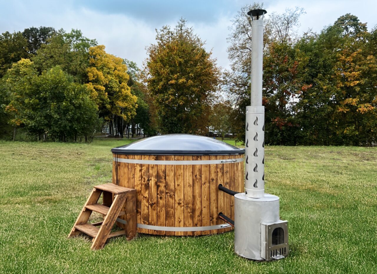 Makoya Spa hot tub DIANA is placed on a green meadow, featuring a wooden cladding, wooden entry steps, an isolating cover and a external metal stove with a tall chimney, sourrounded by a natural landscape. 