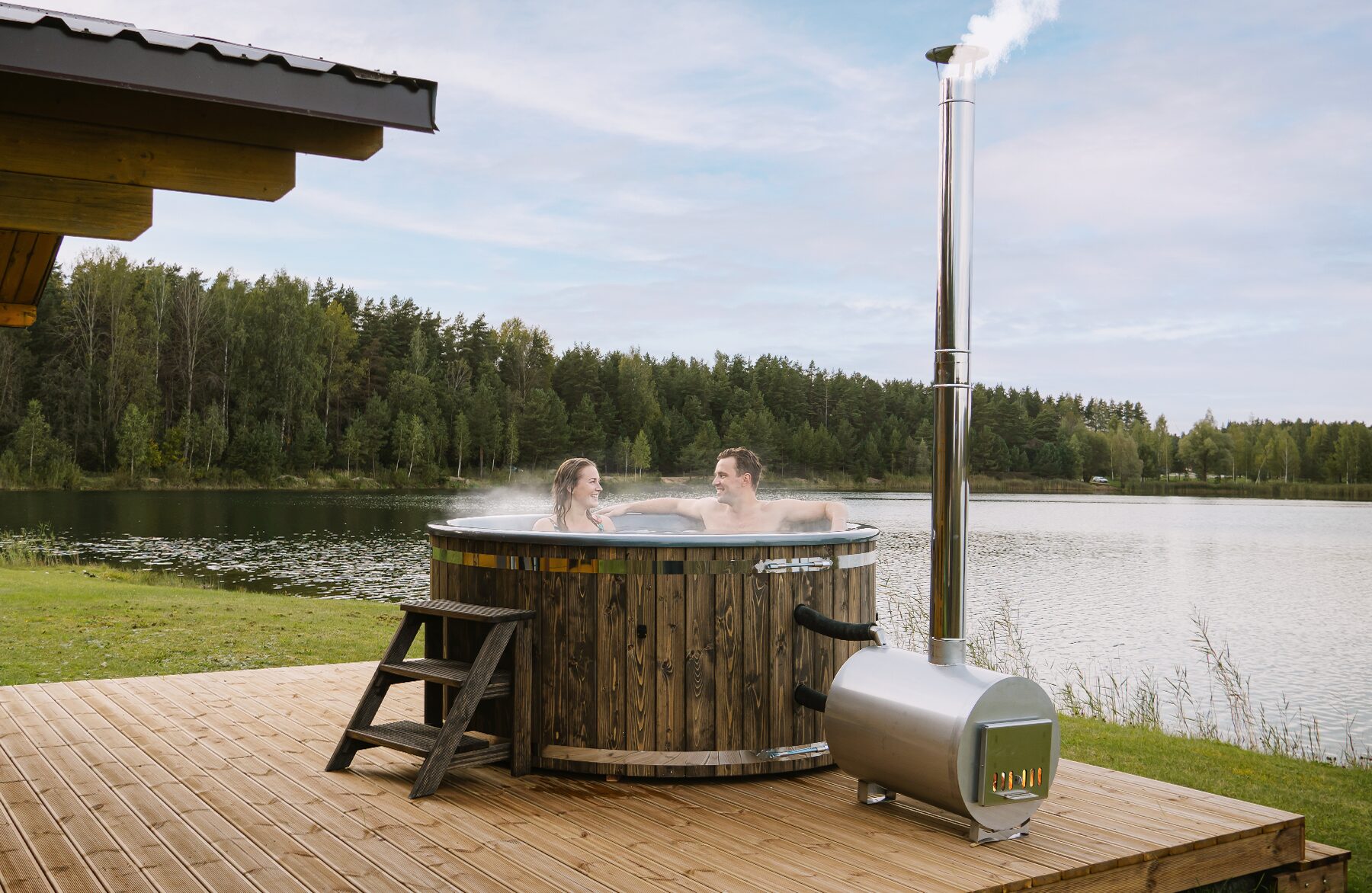 Wooden hot tub LUNA by Makoya Spa on a wooden deck, with external metal stove and chimney, wooden steps and green natural landscape in the background. Two people relax in the steaming hot tub.