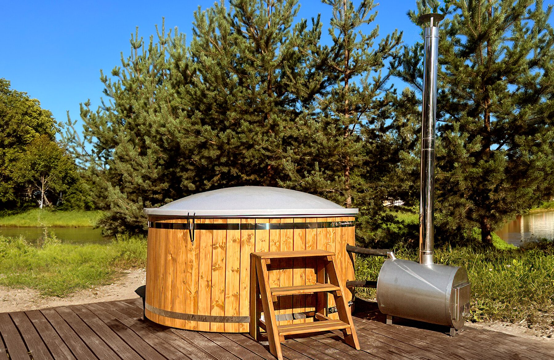 Makoya Spa hot tub LUNA made of wood with a closed cover on a wooden deck, featuring an external metal stove with a tall chimney, wooden entry steps and a green natural landscape in the background.