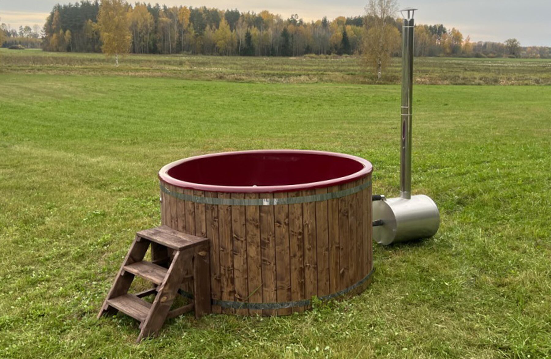 Makoya Spa hot tub LUNA made of wood with a red interior tub on a meadow, featuring an external metal stove with a tall chimney, wooden entry steps and a green natural landscape in the background.