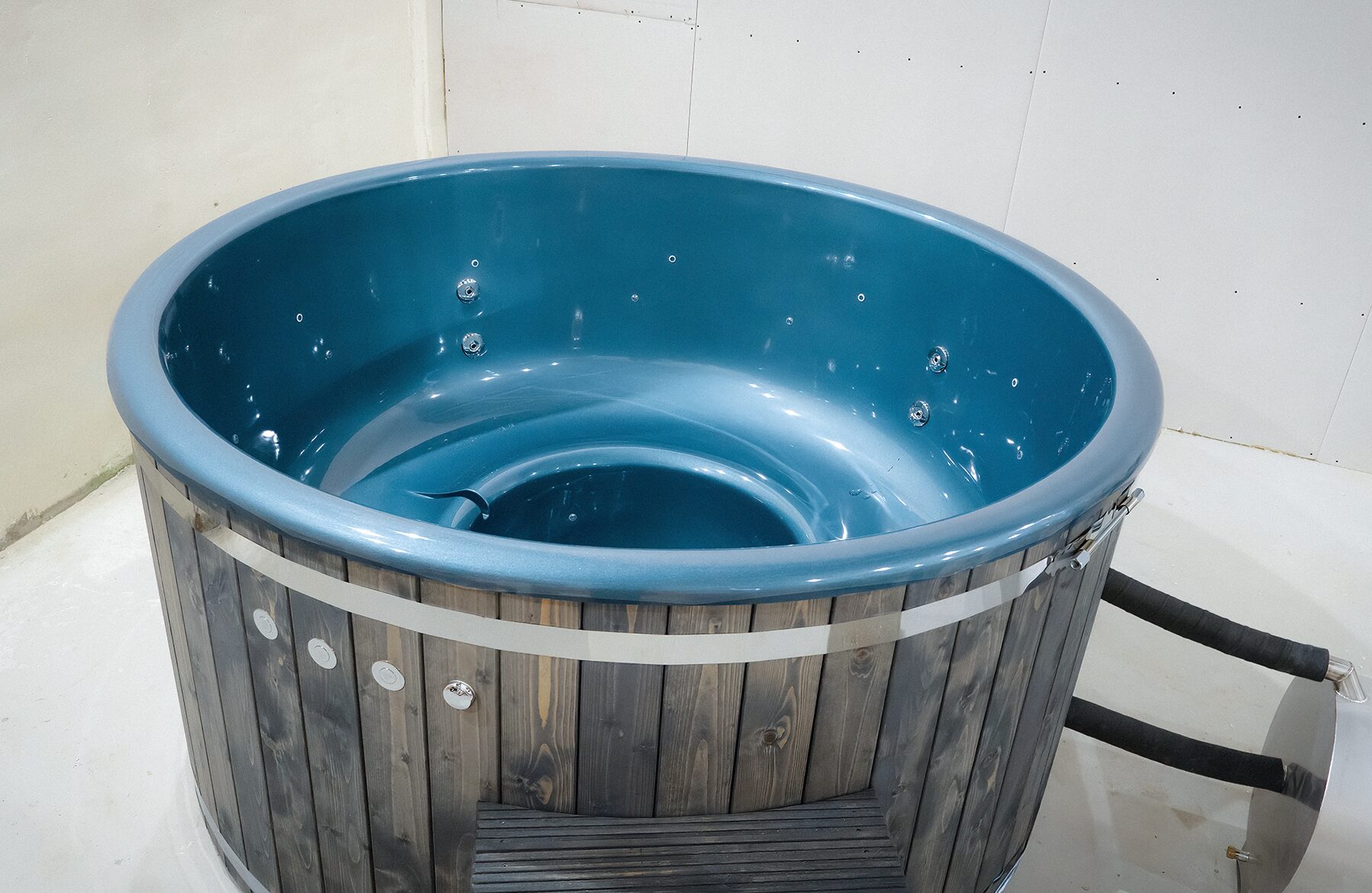 Makoya Spa hot tub LUNA with a blue interior tub and integrated jets, dark wooden cladding, a stainless steel band, is connected to a metal stove.