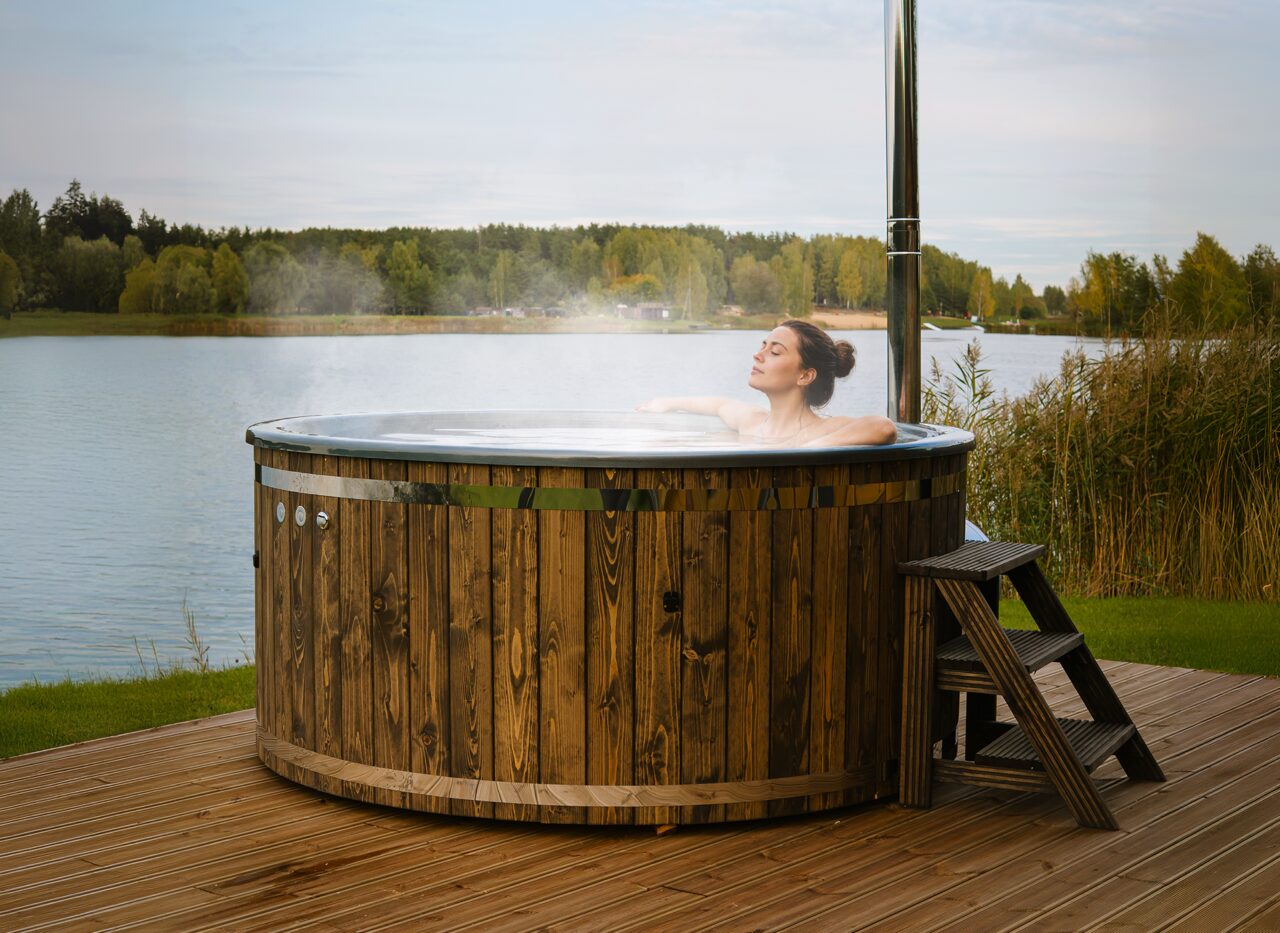 A person relaxing in the warm water of the Makoya Spa hot tub LUNA on a wooden terrace, featuring a metal chimney, entry steps and a calm natural landscape in the backround. 