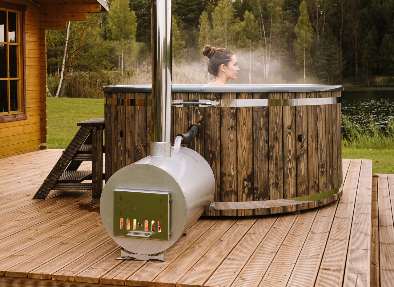 A person relaxing in the Makoya Spa hot tub LUNA on a wooden terrace, featuring a metal stove with chimney, entry steps and landscape in the backround. 