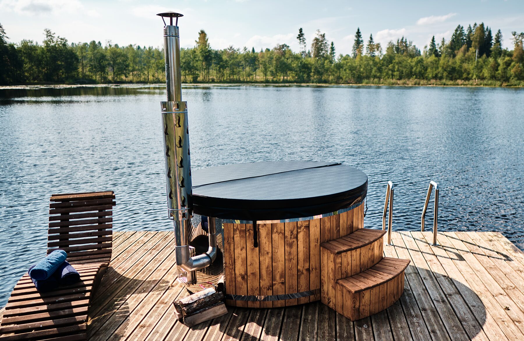 Makoya Spa hot tub SOPHIA with a black cover on a wooden dock by a lake, featuring a metal stove and chimney, steps for entry and a lounge chair, sourrounded by forest.