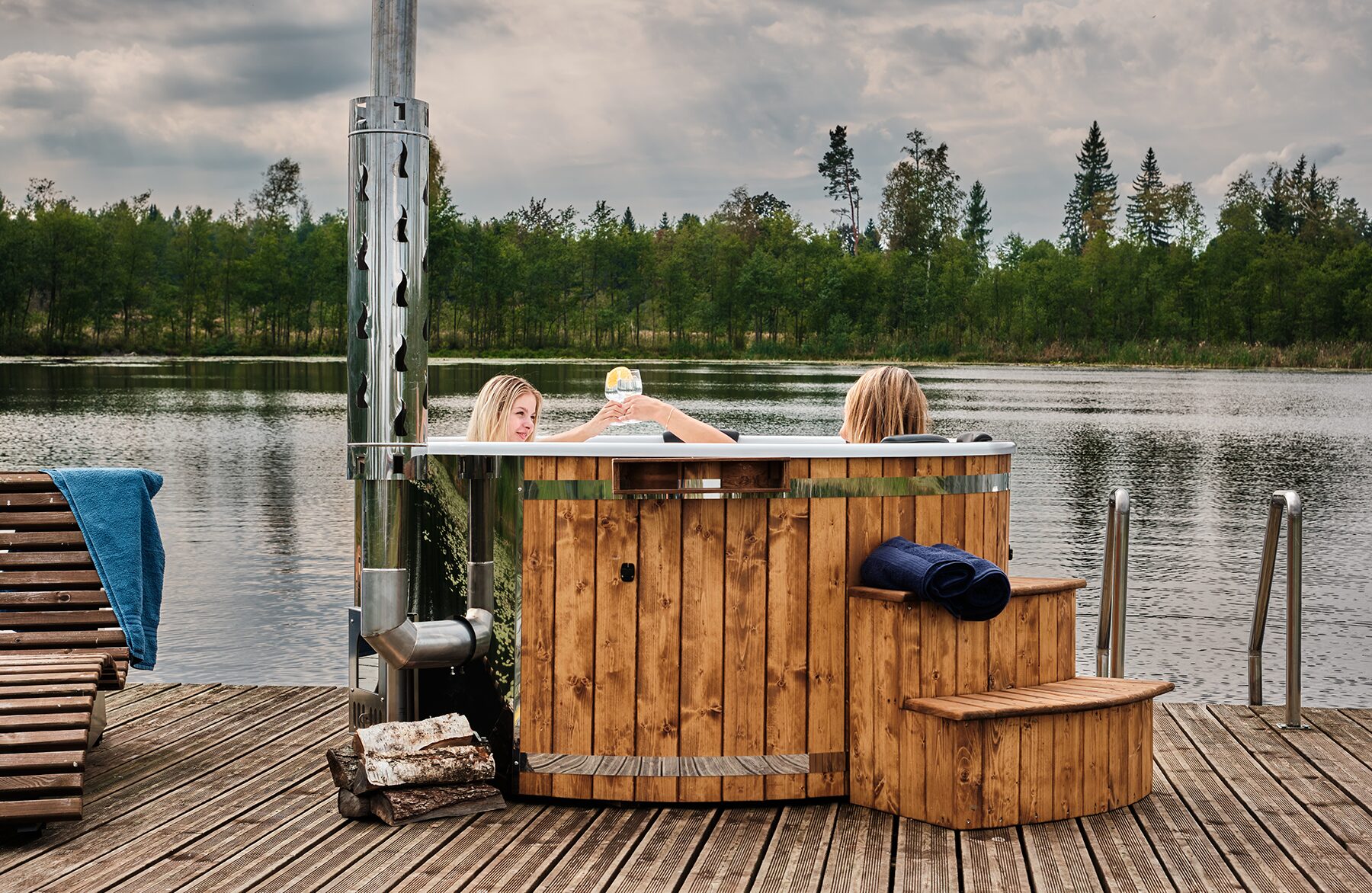 Makoya Spa hot tub SOPHIA, with two people relaxing inside, standing by a lake and featuring a metal stove and chimney, steps for entry and towels, sourrounded by forest.
