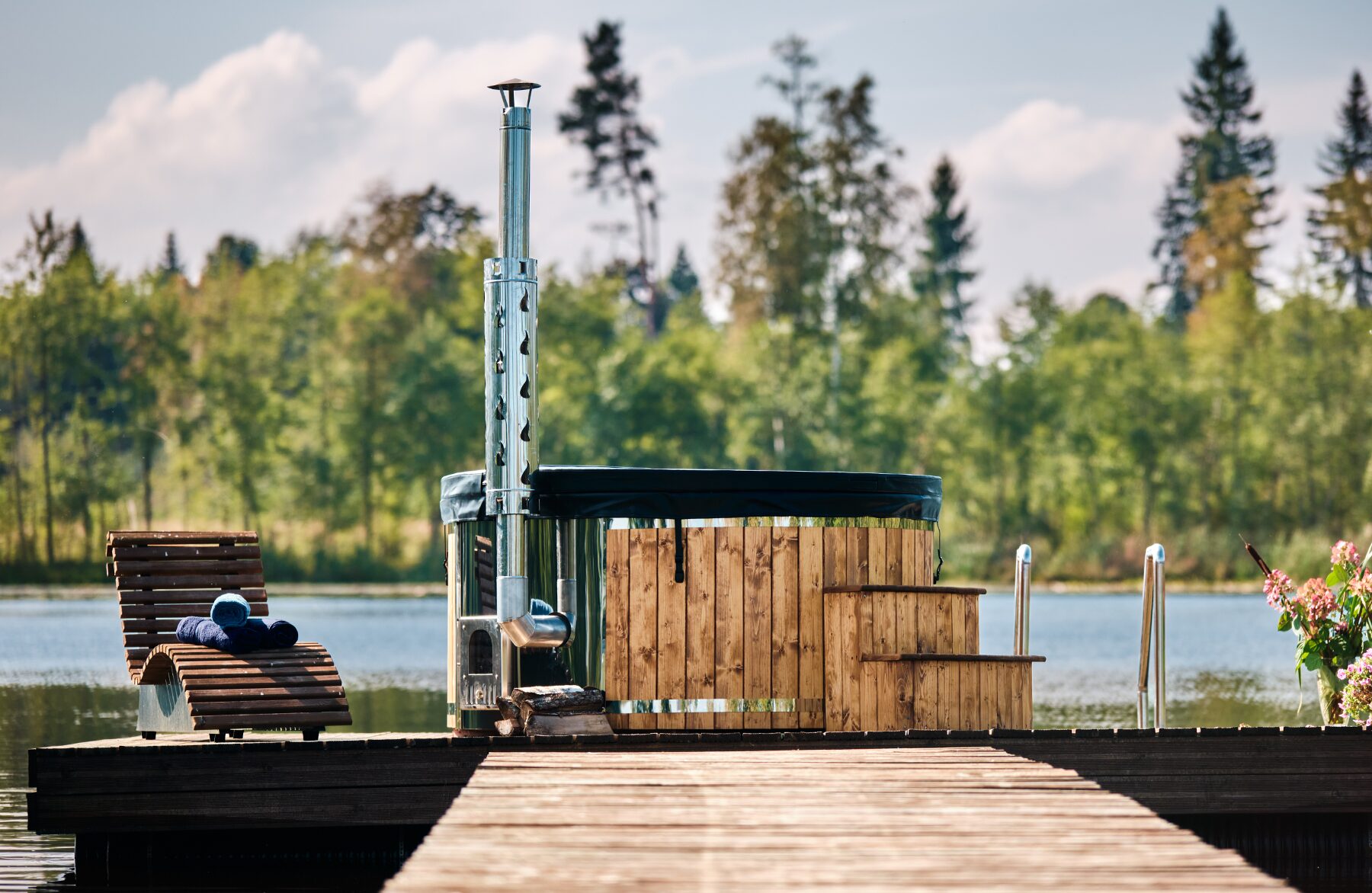 Makoya Spa hot tub SOPHIA with a black cover on a wooden dock by a lake, featuring a stove with a steel stove and chimney, steps for entry and a lounge chair, sourrounded by forest.