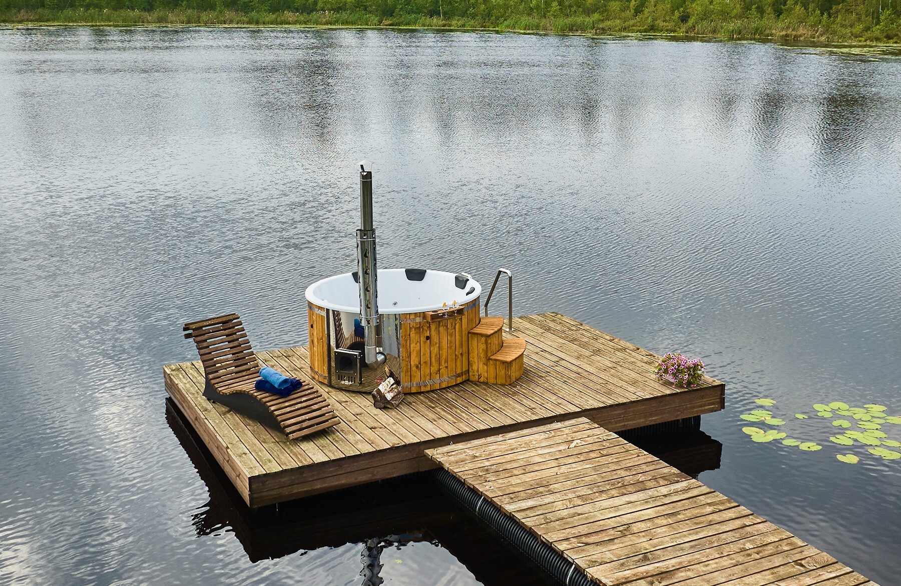 Makoya Spa hot tub SOPHIA with a white interior tub on a wooden dock by a lake, featuring a metal stove and chimney, steps for entry, a lounge chair and towels, sourrounded by nature.