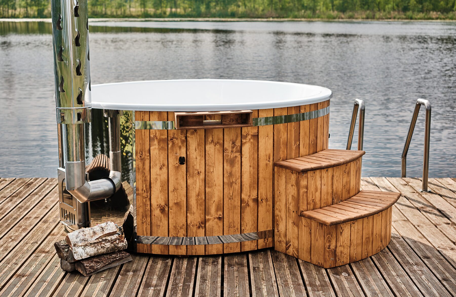 Makoya Spa hot tub SOPHIA with a white interior tub on a wooden dock by a lake, featuring a metal stove and chimney, wooden entry steps and firewood stacked beside it.