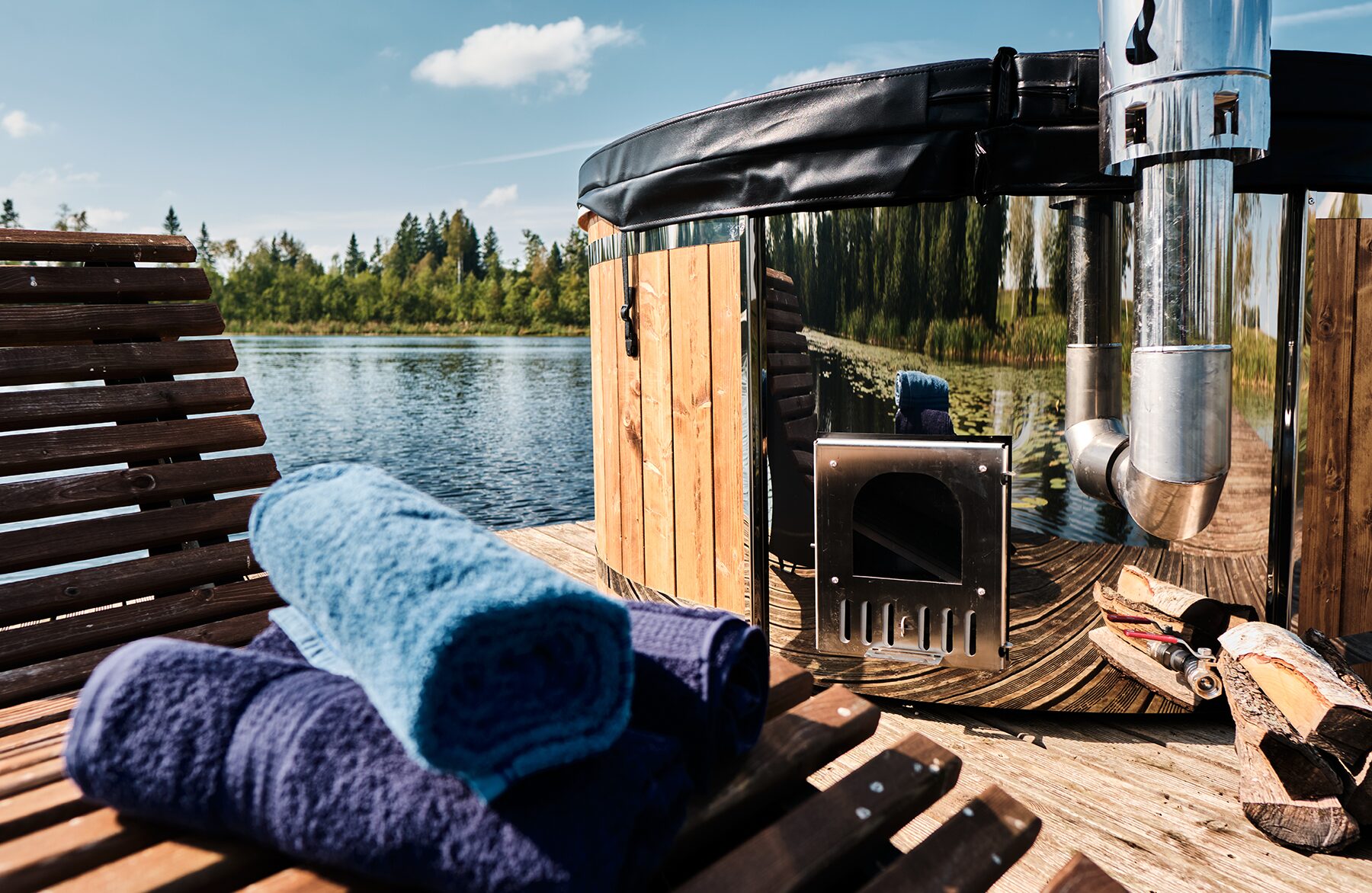 Close-up view Makoya Spa hot tub SOPHIA on a wooden dock by a lake, featuring a black cove, metal stove, and chimney, firewood and neatly folded towels on a wooden lounge chair.