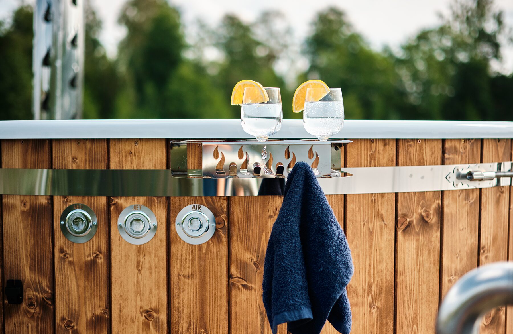 Close-up detail Makoya Spa hot tub SOPHIA featuring a drink holder with two glasses of water and orange slices, a towel holder and wooden cladding set against green background.