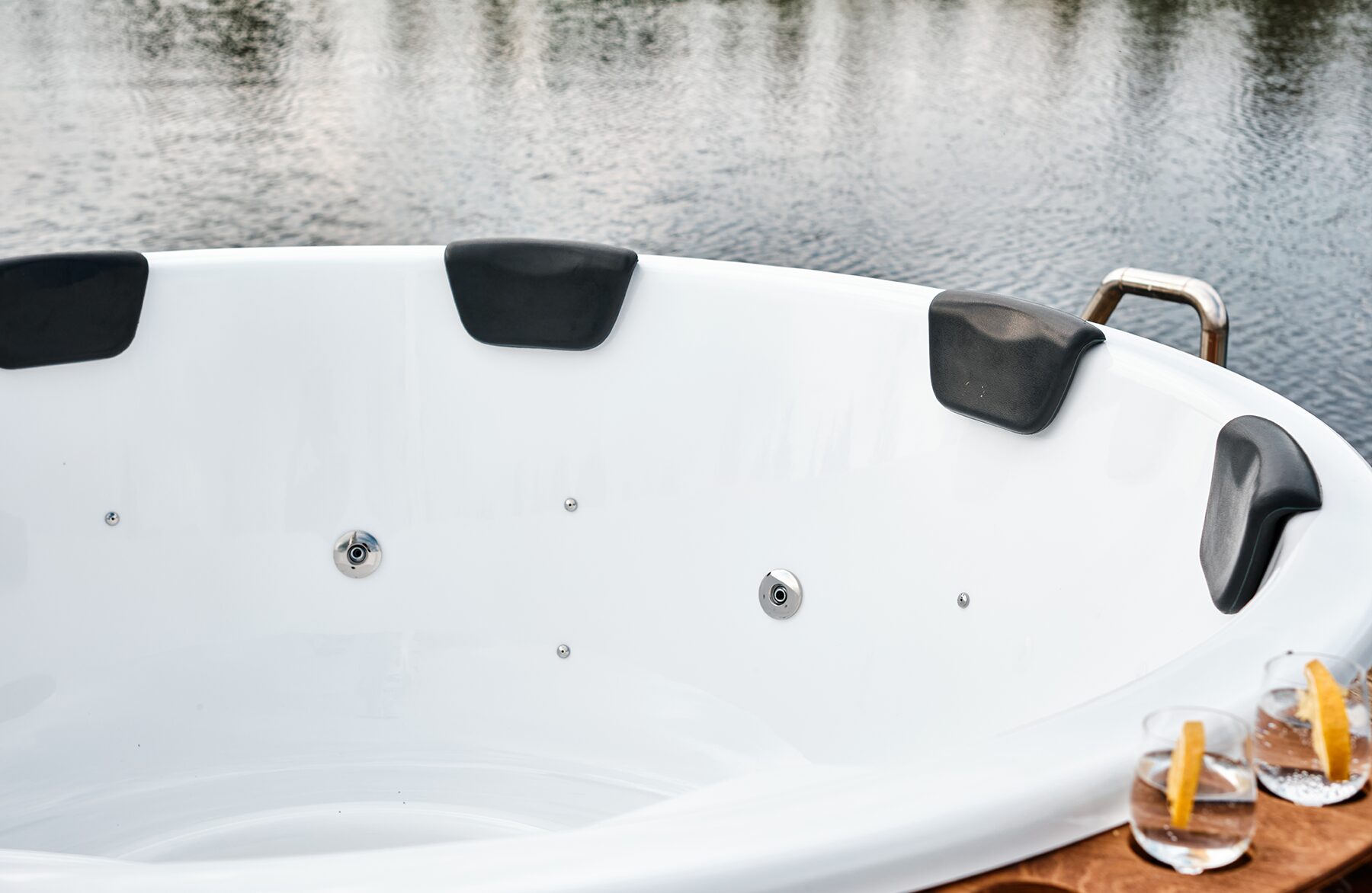 Close-up view Makoya Spa hot tub SOPHIA with a white interior tub with integrated jets and black headrests, a lake can be seen in the backround and drinks placed on the rim.
