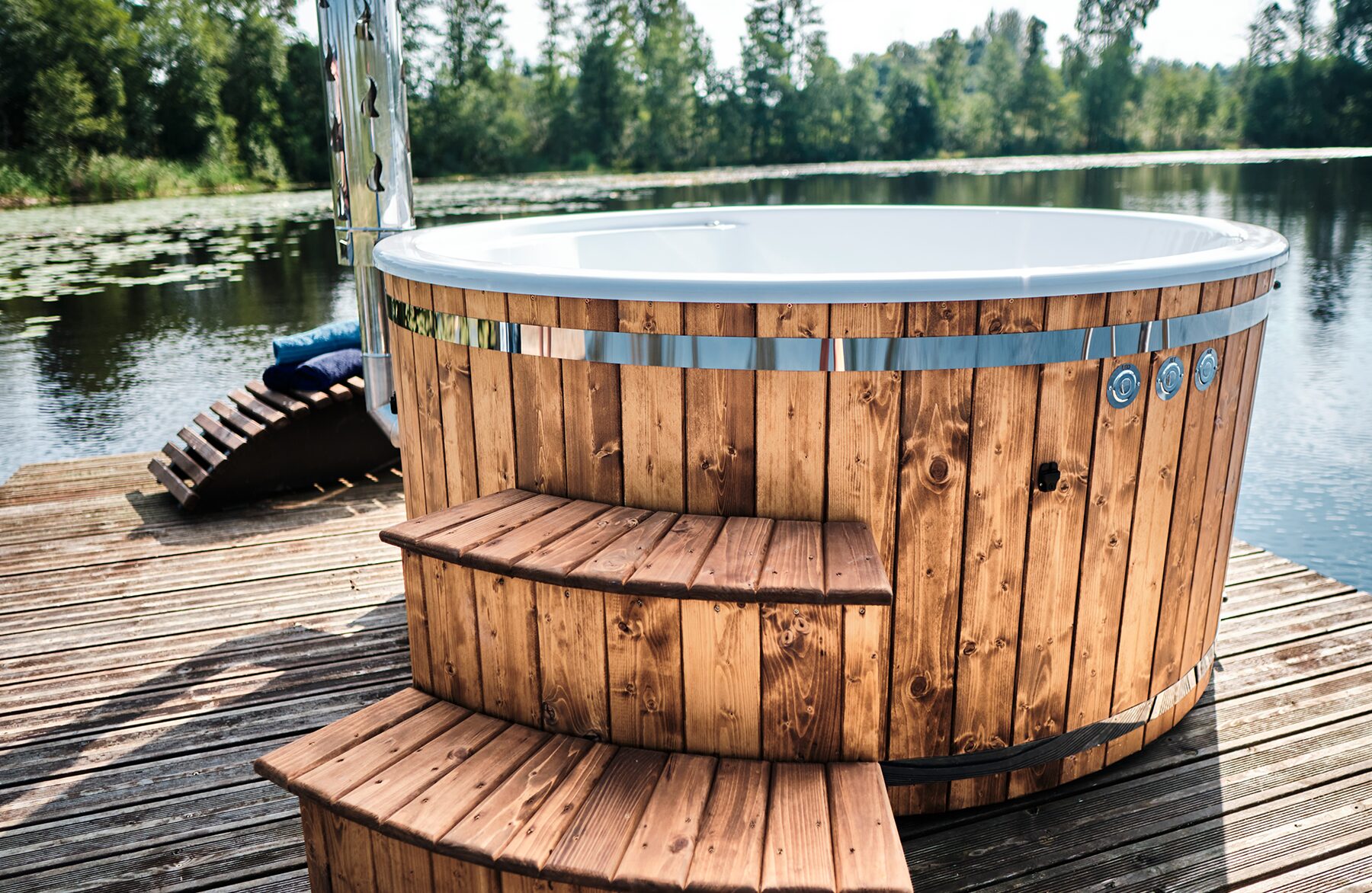 Makoya Spa hot tub SOPHIA with a white interior tub on a wooden dock by a lake, featuring wooden entry steps, a stainless steel band, and a forest landscape in the backround.