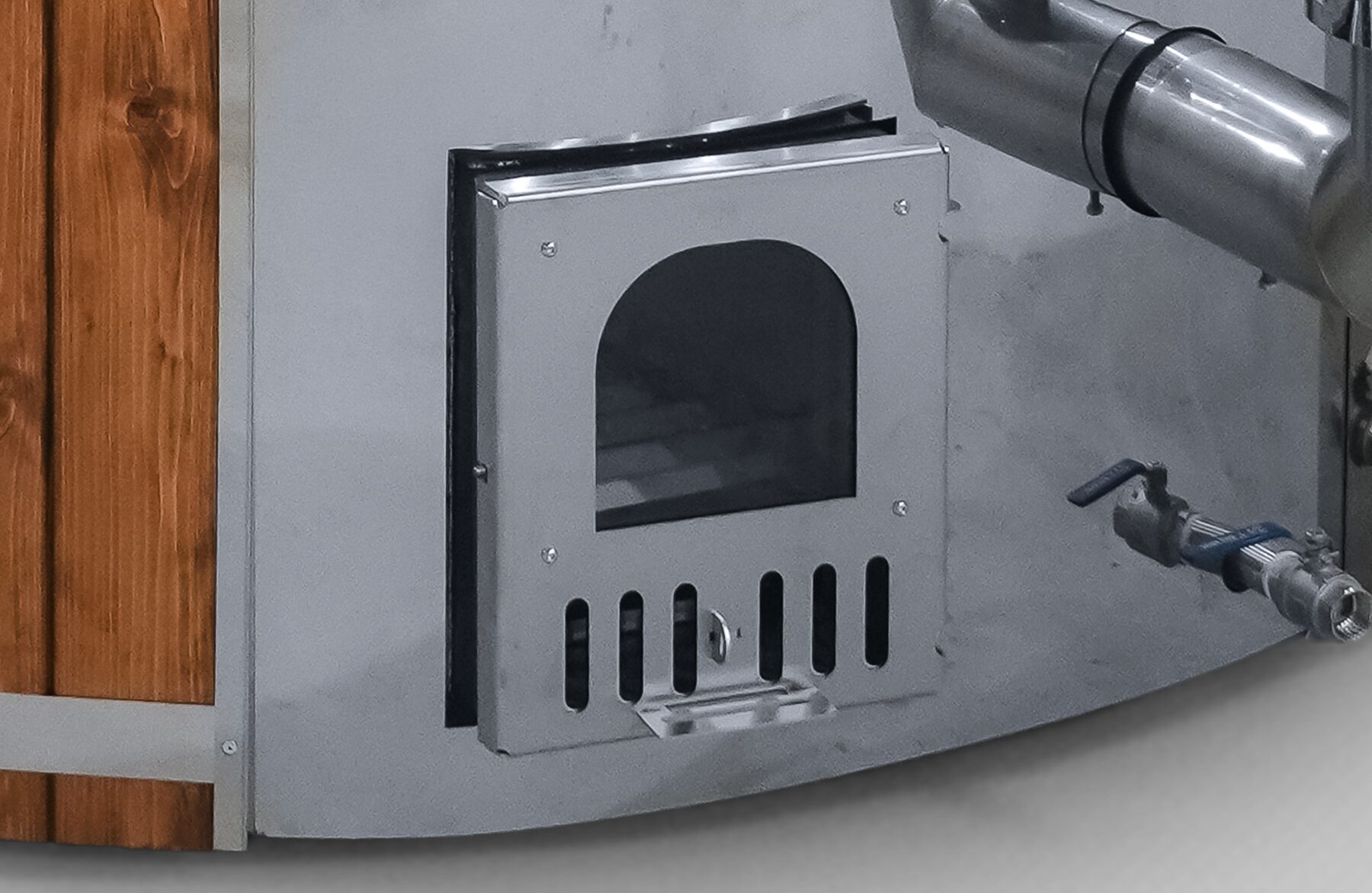 Close-up view of the metal stove of a Makoya Spa hot tub SOPHIA, showing the firebox door, air vents, pipe connection and water drain valve on the exterior of the tub.