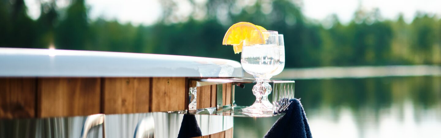 A Makoya Spa hot tub is shown. Two glasses stand on a small shelf and a dark towel hangs from it. In the background a lake surrounded by trees can be seen.