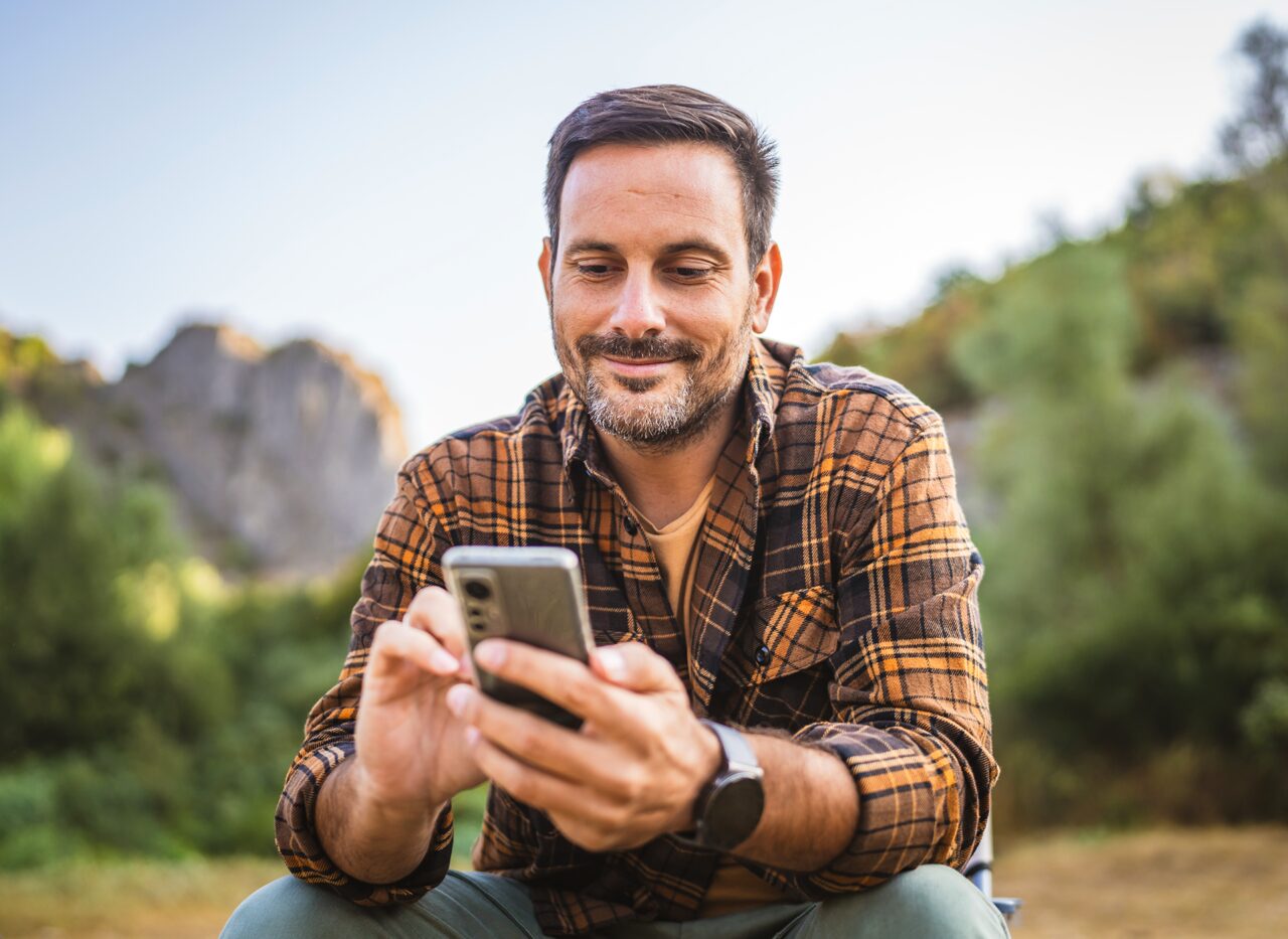 A person is sitting outside, smiling while looking at a smartphone.