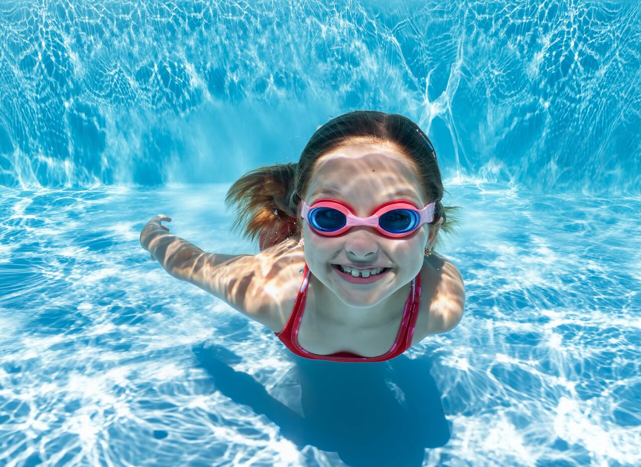 A child smiling while swimming underwater in a clear blue pool, wearing swim goggles, with shimmering light reflections on the water surface and pool floor.