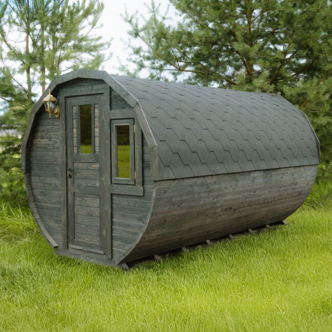 Exterior view of the Makoya Spa tiny house JUNO featuring dark wooden cladding and a curved design, equipped with an entrance door, side window, and shingle roof, set on a green meadow and surrounded by trees.