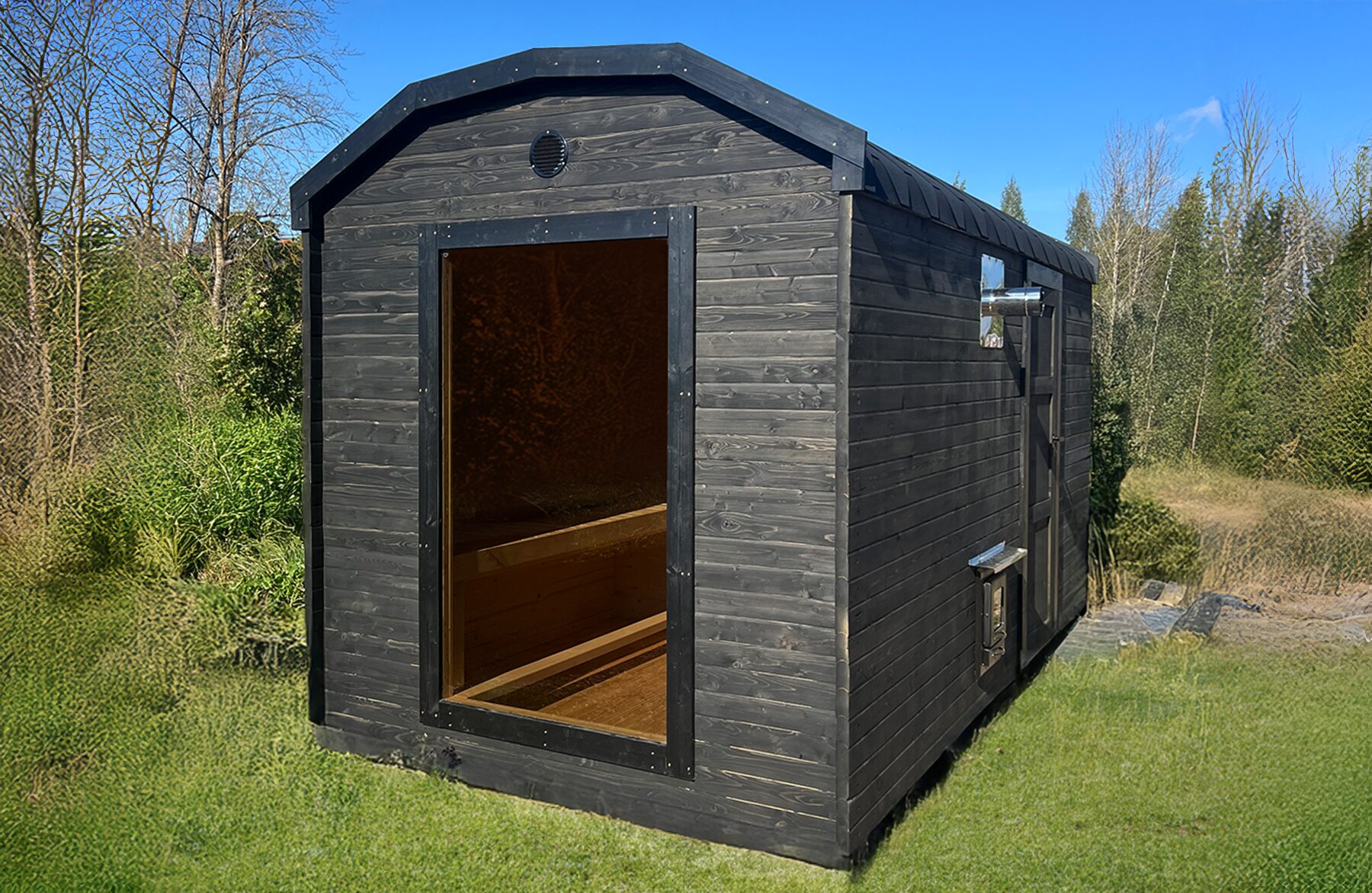Makoya Spa sauna CERES with dark wooden cladding and a glass door is set on a meadow, surrounded by bushes and trees, under blue sky.