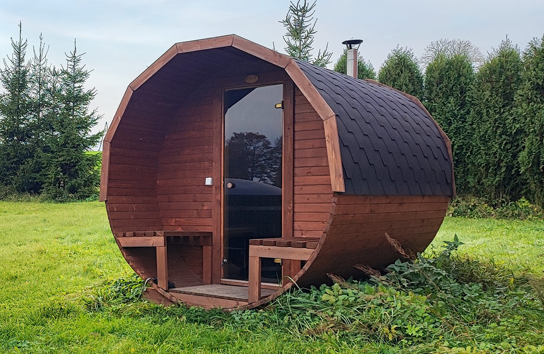 Barrel sauna JUNO by Makoya Spa, on a meadow with trees and blue sky in the background.