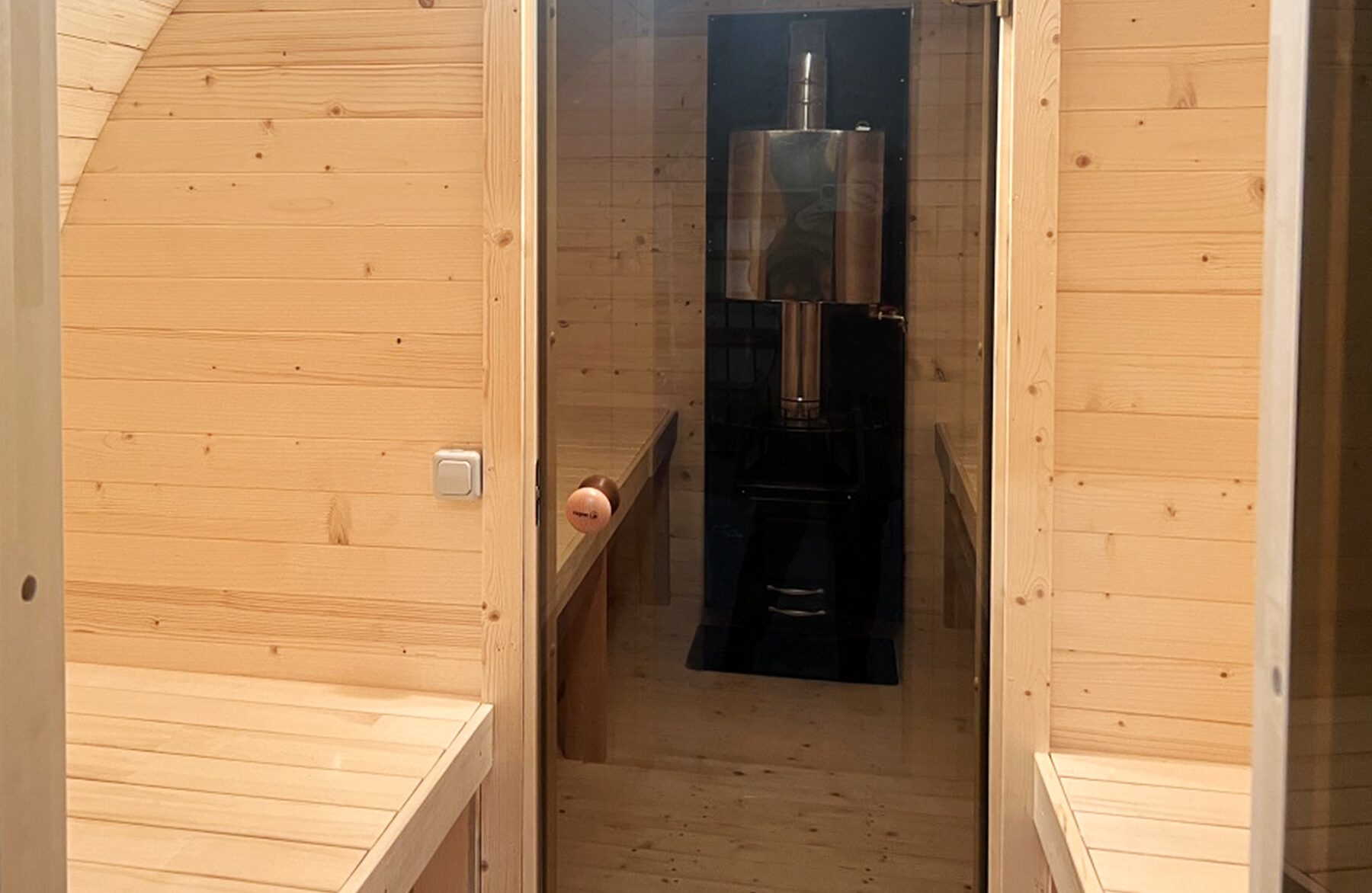 Interiorview of the Makoya Spa barrel sauna JUNO featuring a light wood cladding, facing wooden benches and a view through the glass door toward the integrated sauna stove in the anteroom.