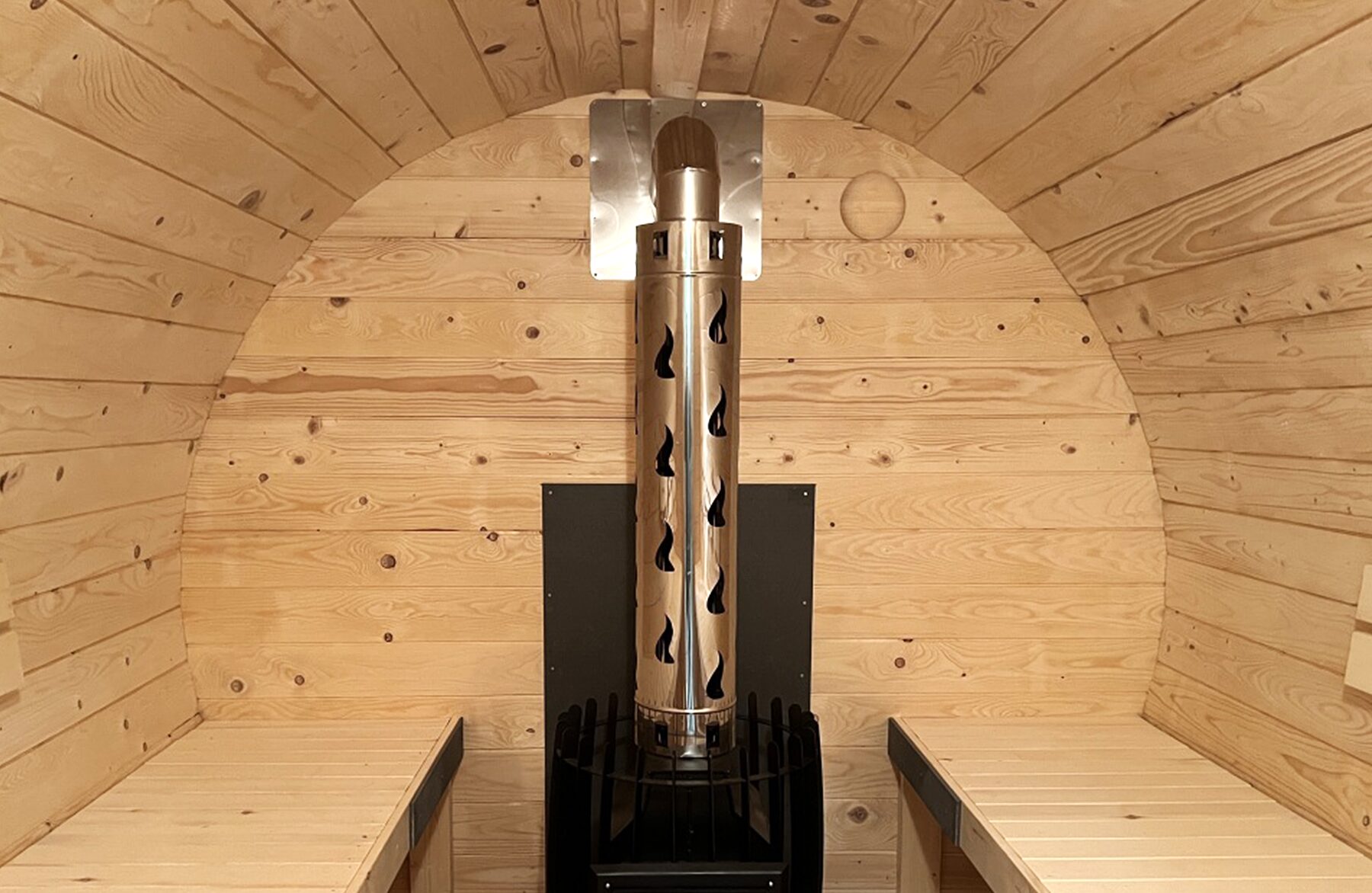 Central interior view of the Makoya Spa barrel sauna JUNO featuring an integrated wood-burning strove with a chimney, surrounded by light wooden cladding and facing benches.