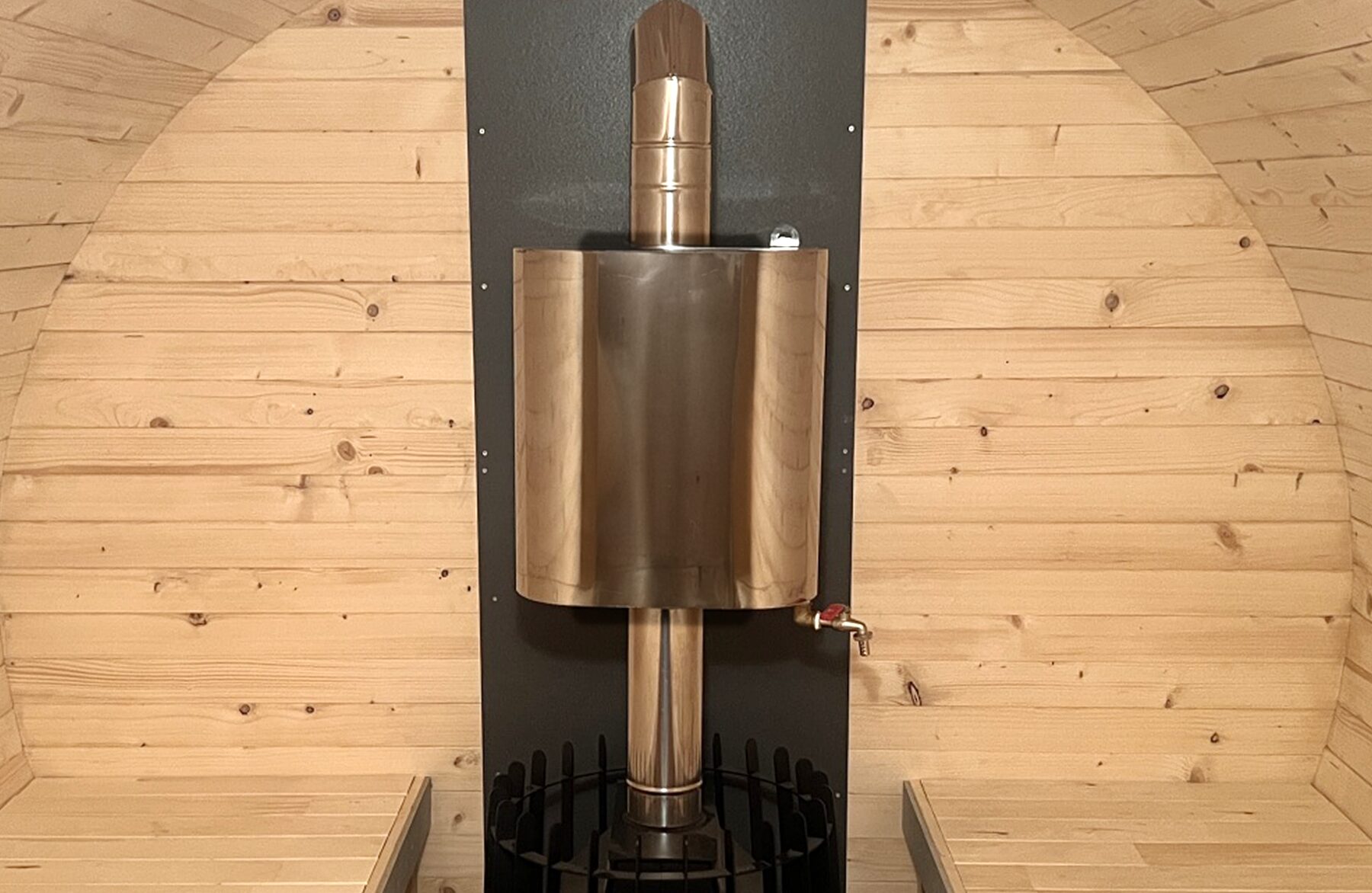 Detailed view of the integrated wood-burning stove with a water tank and stainless steel flue inside the Makoya Spa barrel sauna JUNO, mounted against a dark heat shield and surrounded by light wooden cladding with benches on both sides.