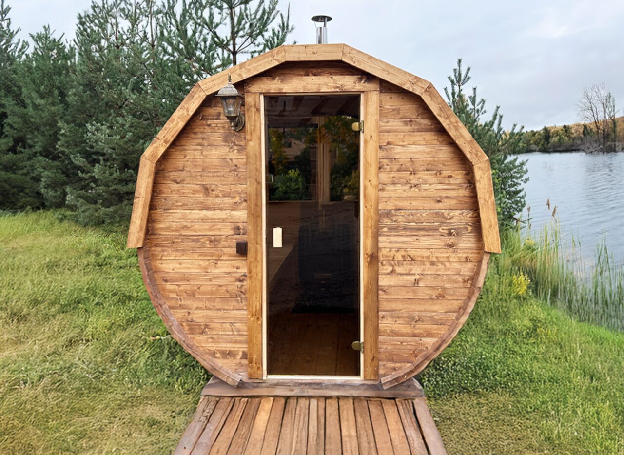 Front view of the Makoya Spa Sauna JUNO featuring a round wooden design, glass door, dark shingle roof and a wooden deck, set on a green meadow by a lake surrounded by trees and a peaceful landscape.