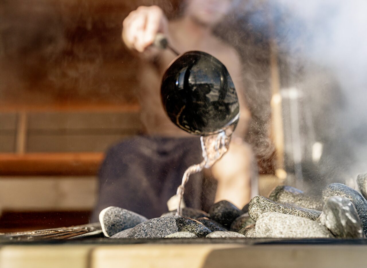 A Person pours water with a ladle over hot sauna stones inside the Makoya Spa Sauna JUNO, creating rising steam.