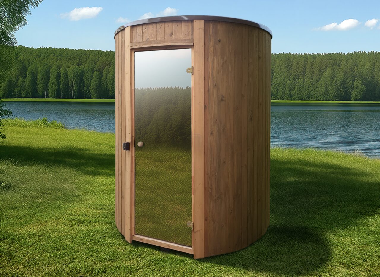 Exterior view of the sauna VESTA by Makoya Spa featuring light wooden cladding and a glass door in a round design, set on a green meadow directly by a lake, surrounded by nature.