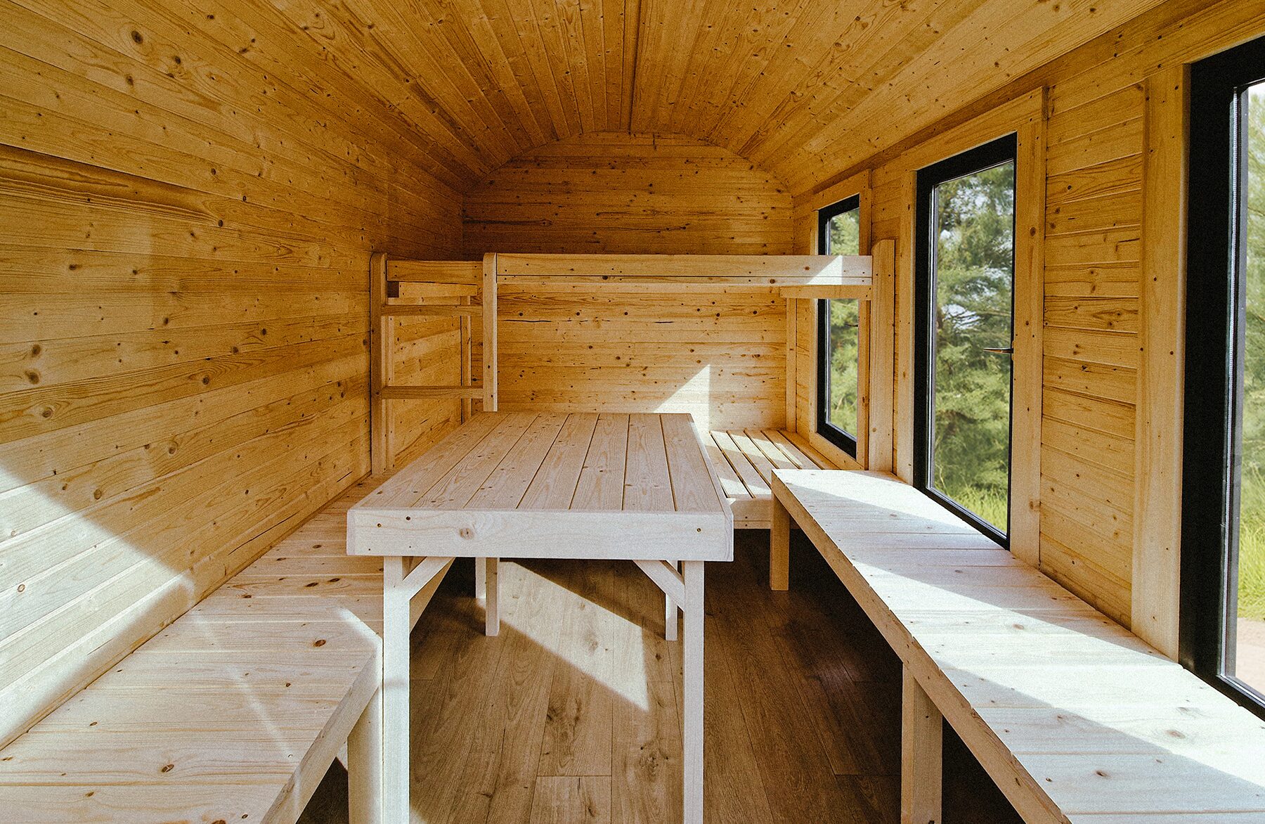 Interior view of a Makoya Spa tiny house CERES featuring light wooden paneling and a gently curved ceiling. The space is open and functional, with built-in benches along the walls and a solid table in the center. Large windows flood the interior with natural light and offer views of the surrounding greenery, creating a warm and natural living atmosphere.
