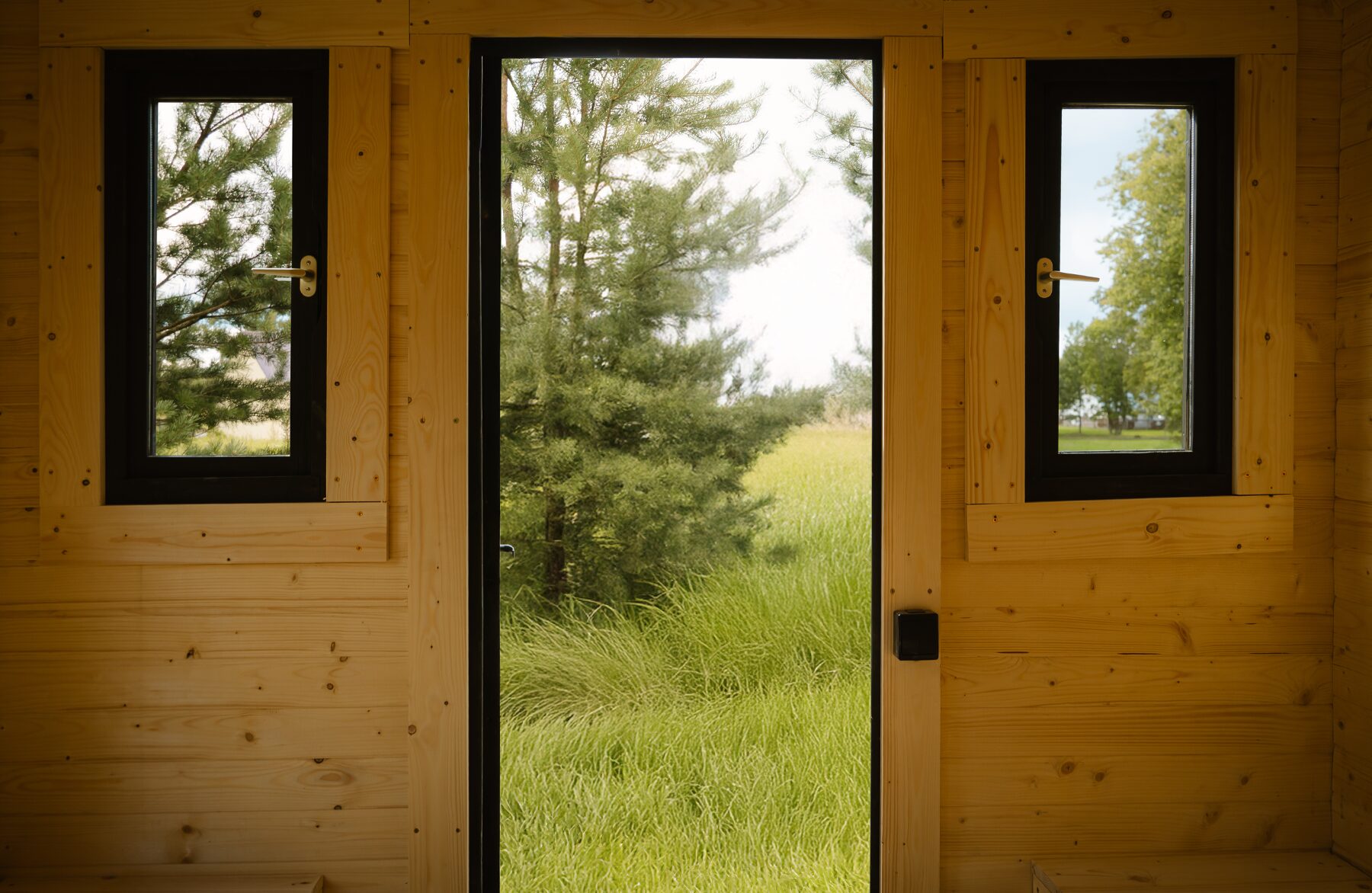 Interior view of a Makoya Spa tiny house CERES featuring a central entrance door flanked by two narrow windows. The light wooden interior creates a warm and natural atmosphere, while the view through the door and windows opens onto grass and trees, emphasizing a close connection to the surrounding landscape.