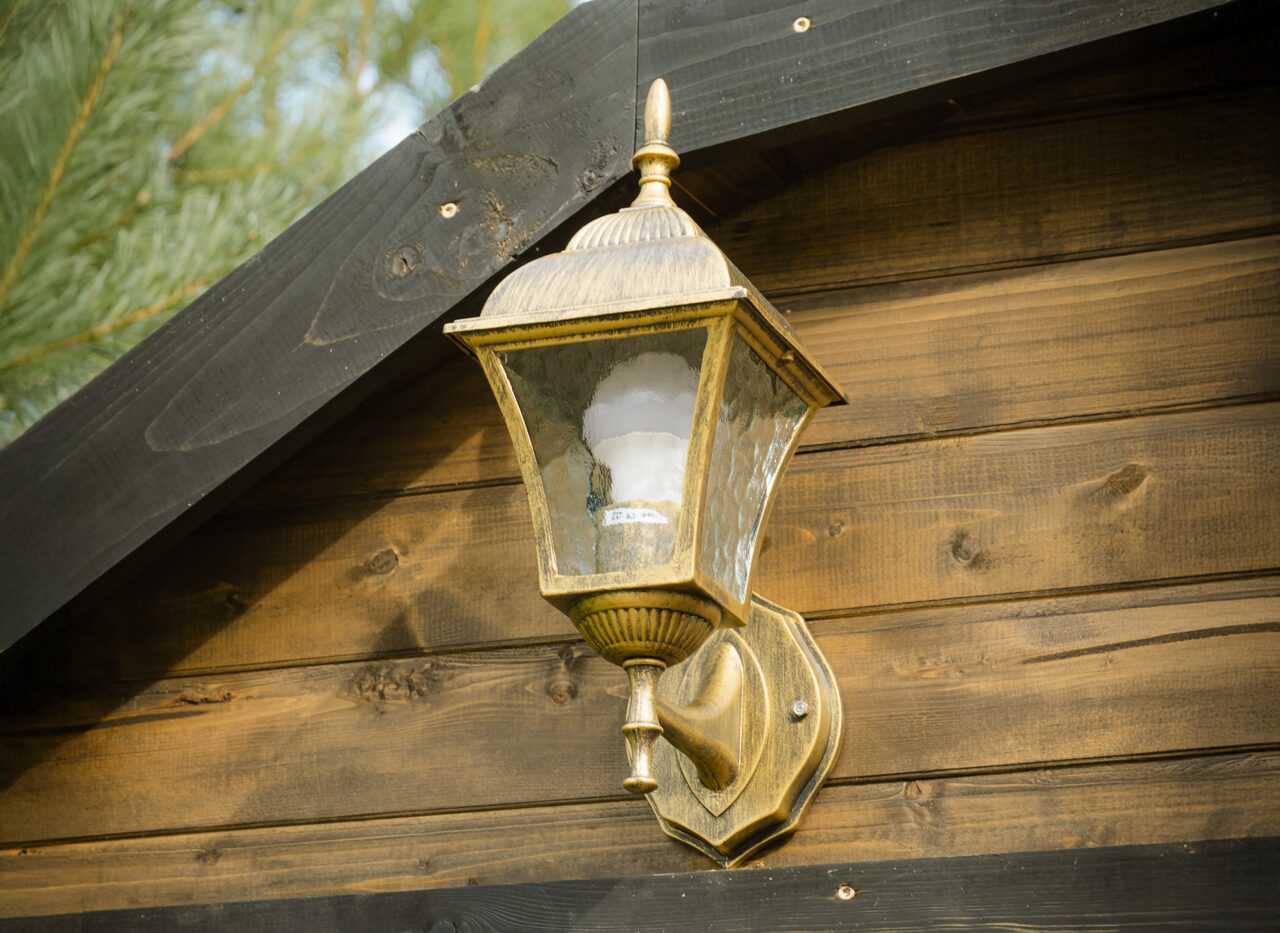 Close-up view of a classic metal-and-glass outdoor wall lantern mounted on the wooden facade of a Makoya Spa tiny house CERES. The light is positioned above the entrance and complements the rustic appearance of the dark wooden exterior.