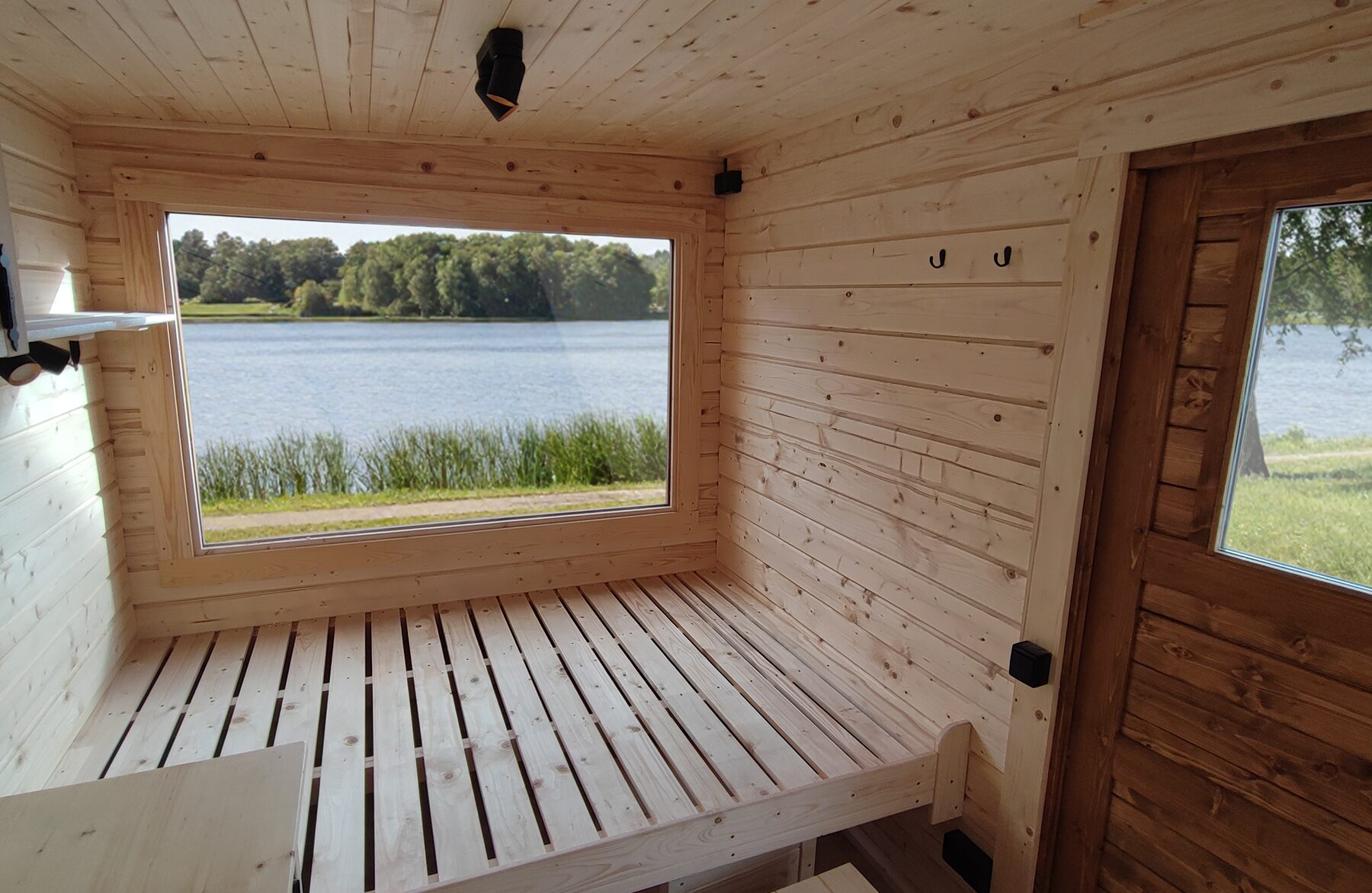 Interior view of the Makoya Spa tiny house DELUXE featuring a light wooden interior, a spacious sleeping area, and a large panoramic window overlooking a lake and reeds, filled with natural light and a close-to-nature atmosphere.