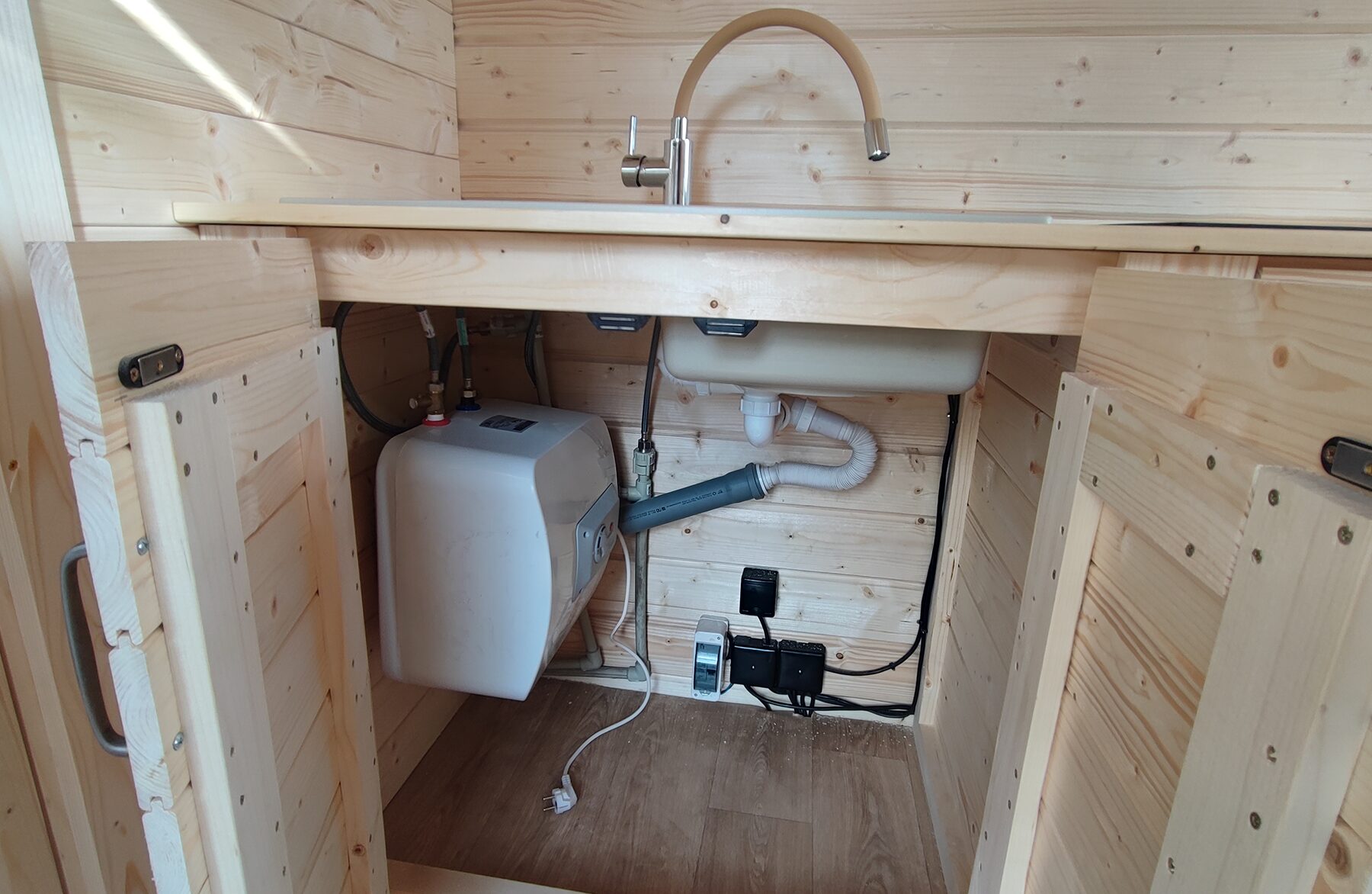 Detailed view of the kitchen area in a Makoya Spa tiny house DELUXE with open base cabinet doors: visible are the sink with faucet, the water installation including a water heater, drainage pipes, and electrical connections. The light wood paneling highlights the functional and space-efficient design.