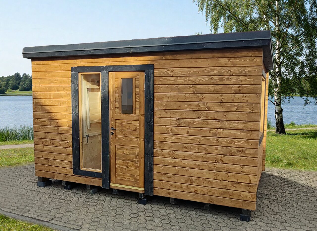 Exterior view of the Makoya Spa tiny house DELUXE with wooden cladding and dark window frames, placed on paved ground by a lake, surrounded by green grass, trees, and a calm natural landscape.