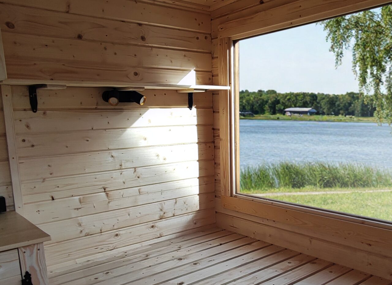 Interior view of the Makoya Spa tiny house DELUXE featuring a light wooden interior, a spacious sleeping area, and a large panoramic window overlooking a lake and reeds, filled with natural light and a close-to-nature atmosphere.