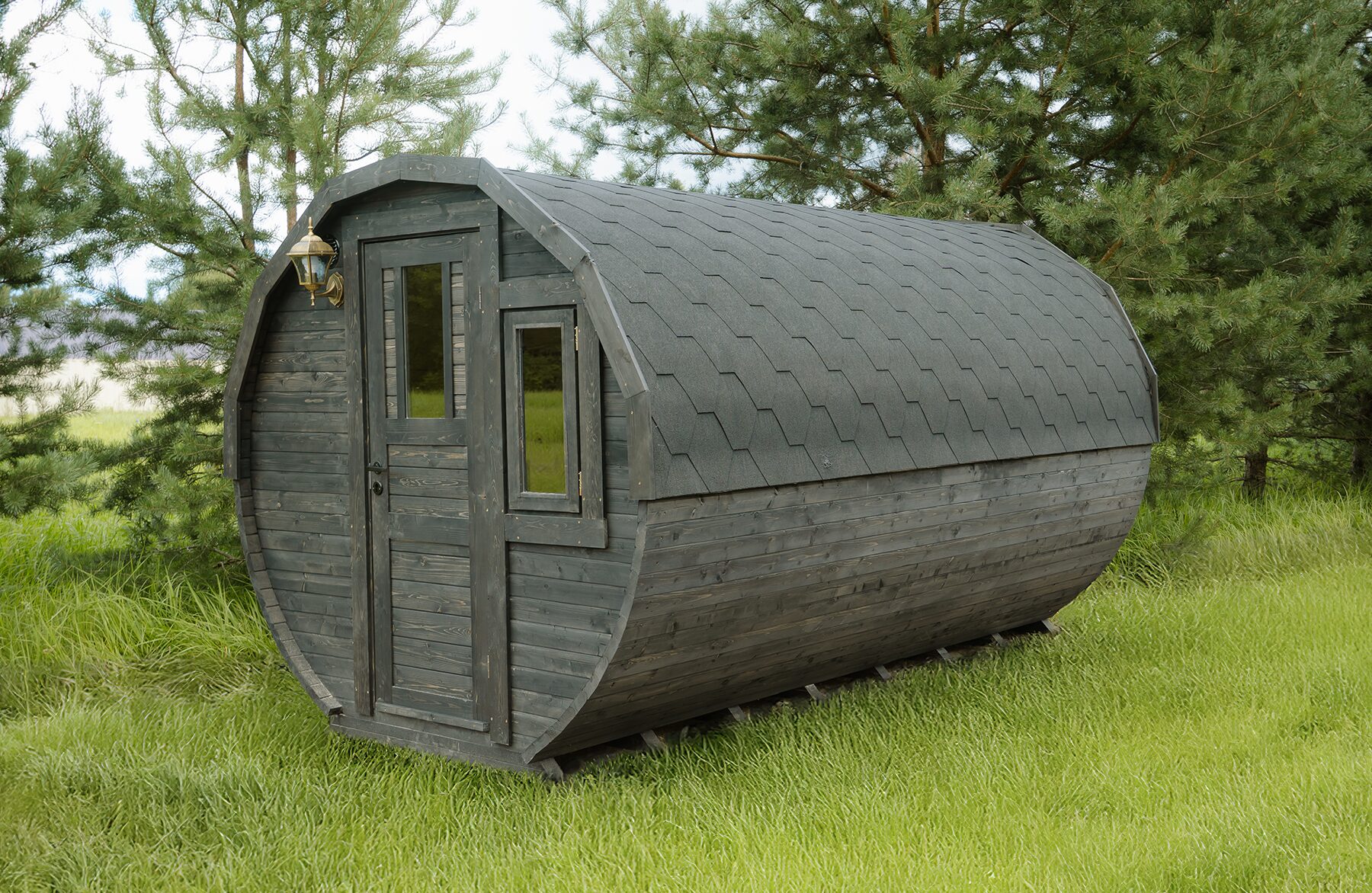 Exterior view of the Makoya Spa tiny house JUNO featuring dark wooden cladding and a curved design, equipped with an entrance door, side window, and shingle roof, set on a green meadow and surrounded by trees.