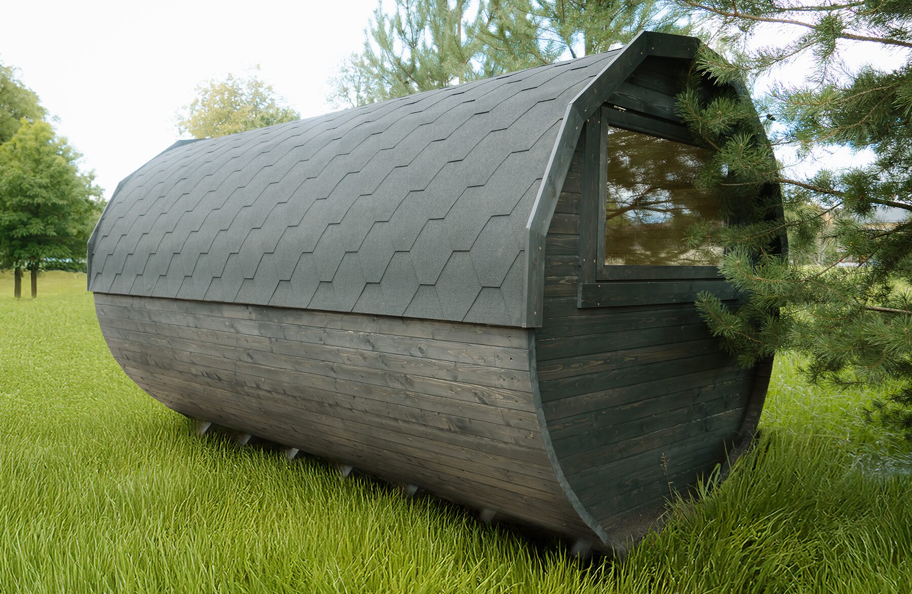 Rear and side view of the Makoya Spa tiny house JUNO with dark wooden cladding and a curved shingle roof, featuring a large window, set on a green meadow and surrounded by trees.