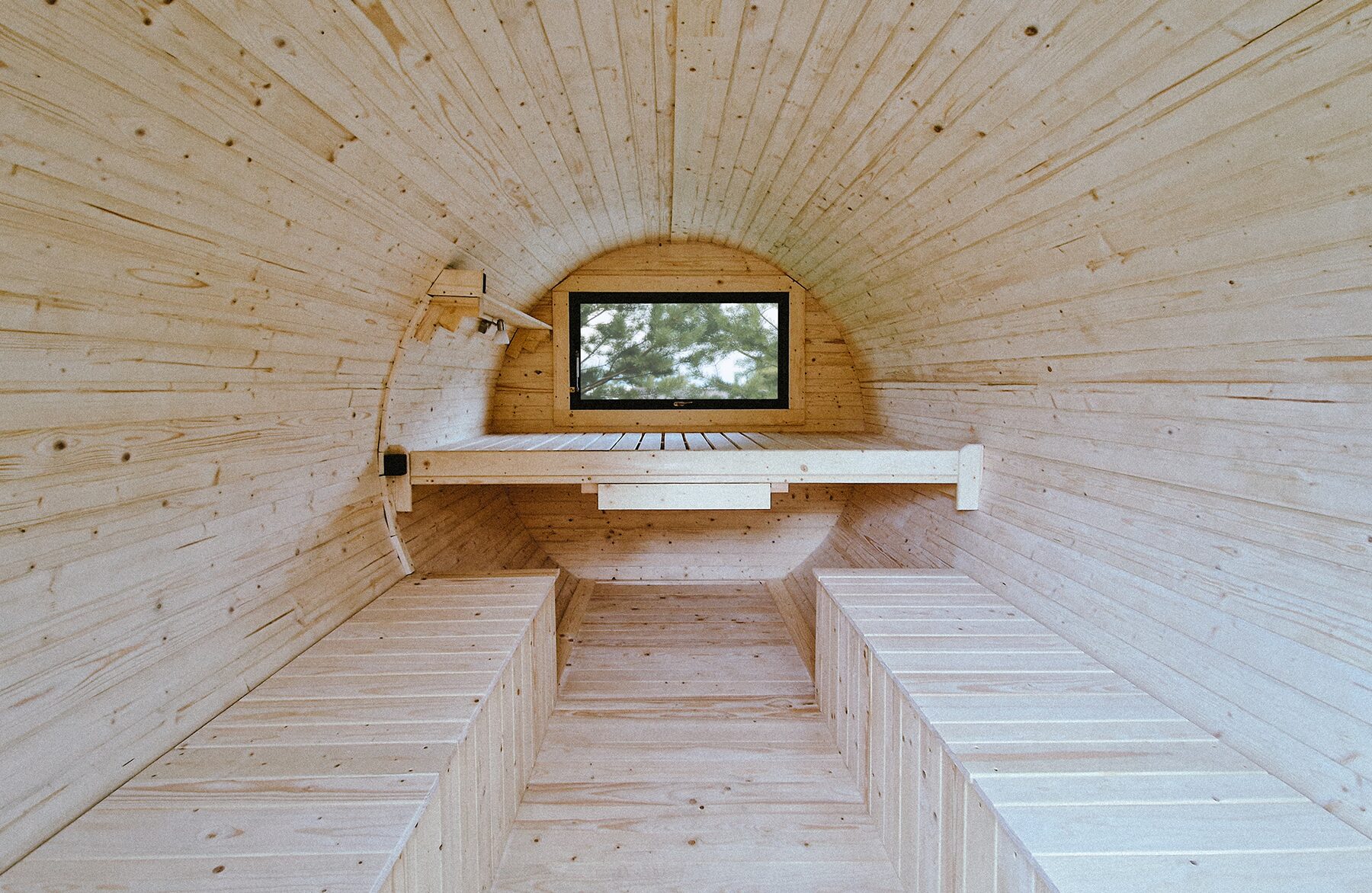Interior view of the Makoya Spa tiny house JUNO featuring light wooden paneling, two opposing benches, and an elevated reclining bench at the rear. A small window provides natural light and a view of the surrounding greenery.