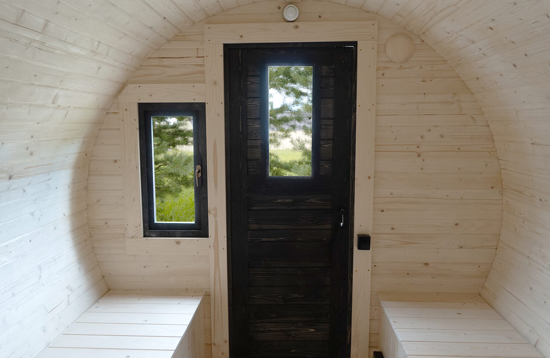 Interior view of the Makoya Spa tiny house JUNO featuring light wooden paneling, a black entrance door with a narrow viewing window, and a side window. Two bright wooden benches on the left and right provide comfortable seating, while natural light and views of greenery create a calm and relaxing atmosphere.