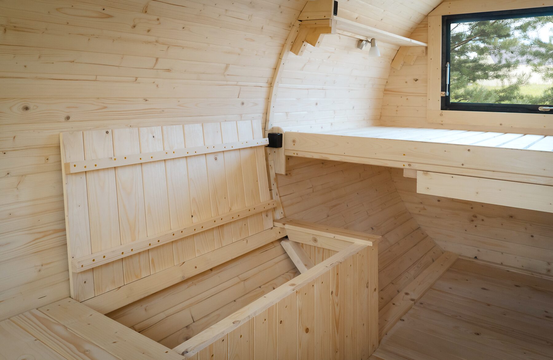 Detailed view of the interior of the Makoya Spa tiny house JUNO with light wood panelling. You can see a raised sleeping area, a fold-out bench and a window with a view of the countryside. The clear wood structure and well-thought-out design create a bright and natural atmosphere.