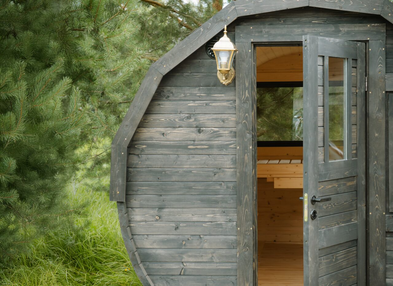 Close-up exterior view of the Makoya Spa tiny house JUNO with dark-stained wooden cladding. The open door reveals the bright wooden interior with benches inside. A classic outdoor lantern is mounted on the facade, and the tiny house is set in lush grass surrounded by pine trees.