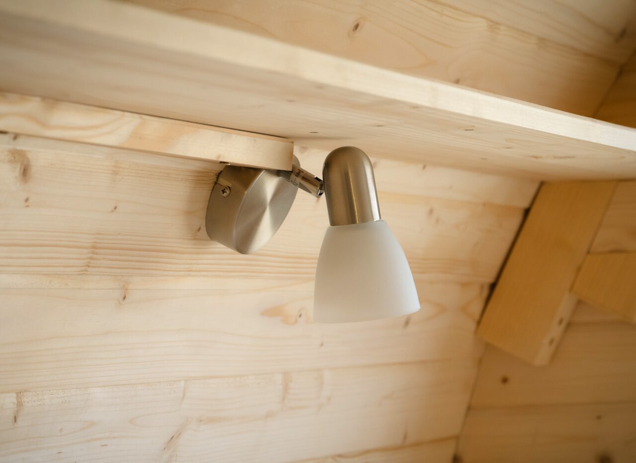 Close-up of the interior lighting of the Makoya Spa tiny house JUNO. A modern, adjustable wall-mounted lamp made of metal with a frosted glass shade is installed on the light wooden paneling, providing warm, indirect light inside the tiny house.