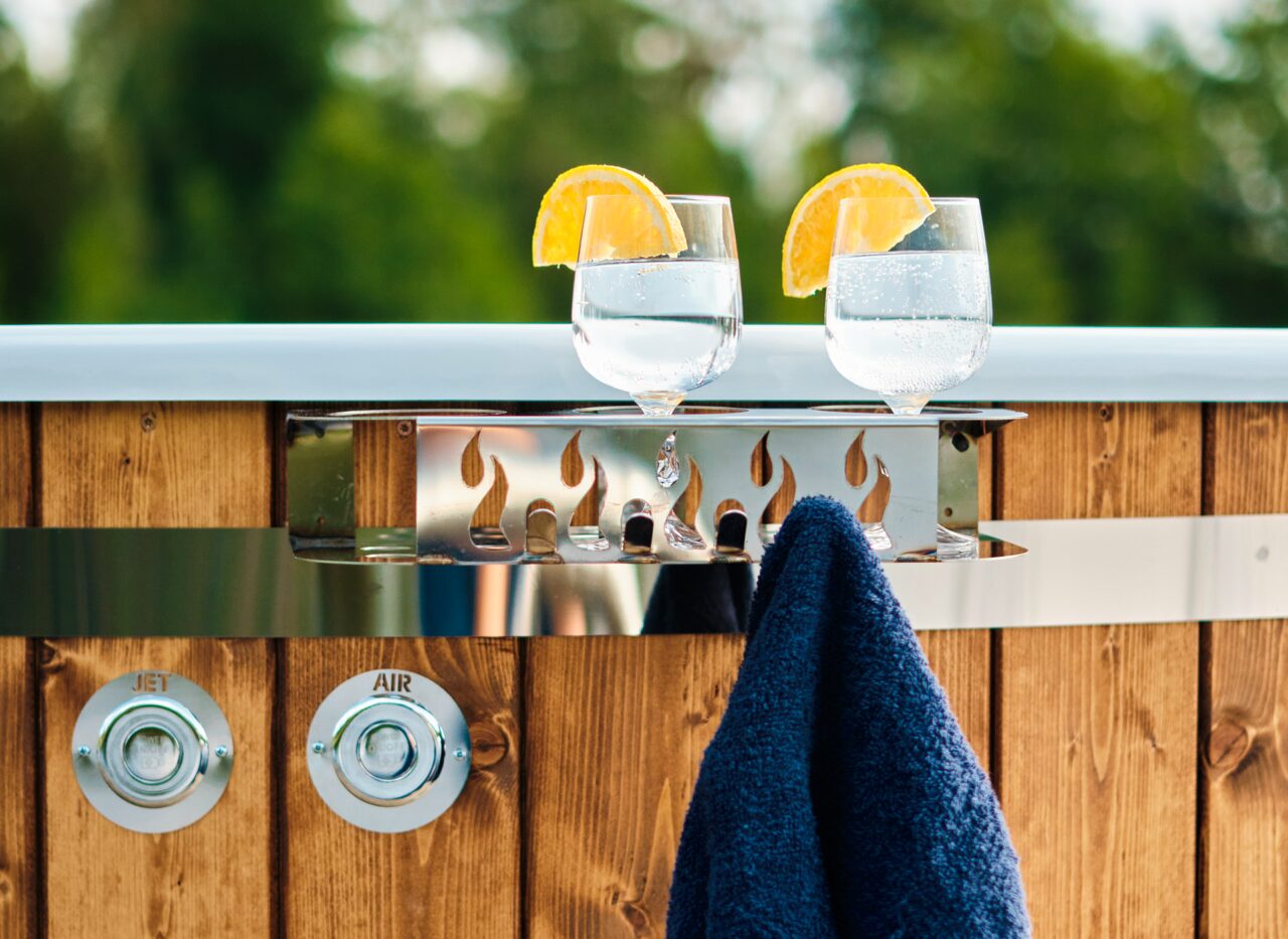A Makoya Spa hot tub is shown. Two glasses stand on a small shelf and a dark towel hangs from it. In the background trees can be seen.