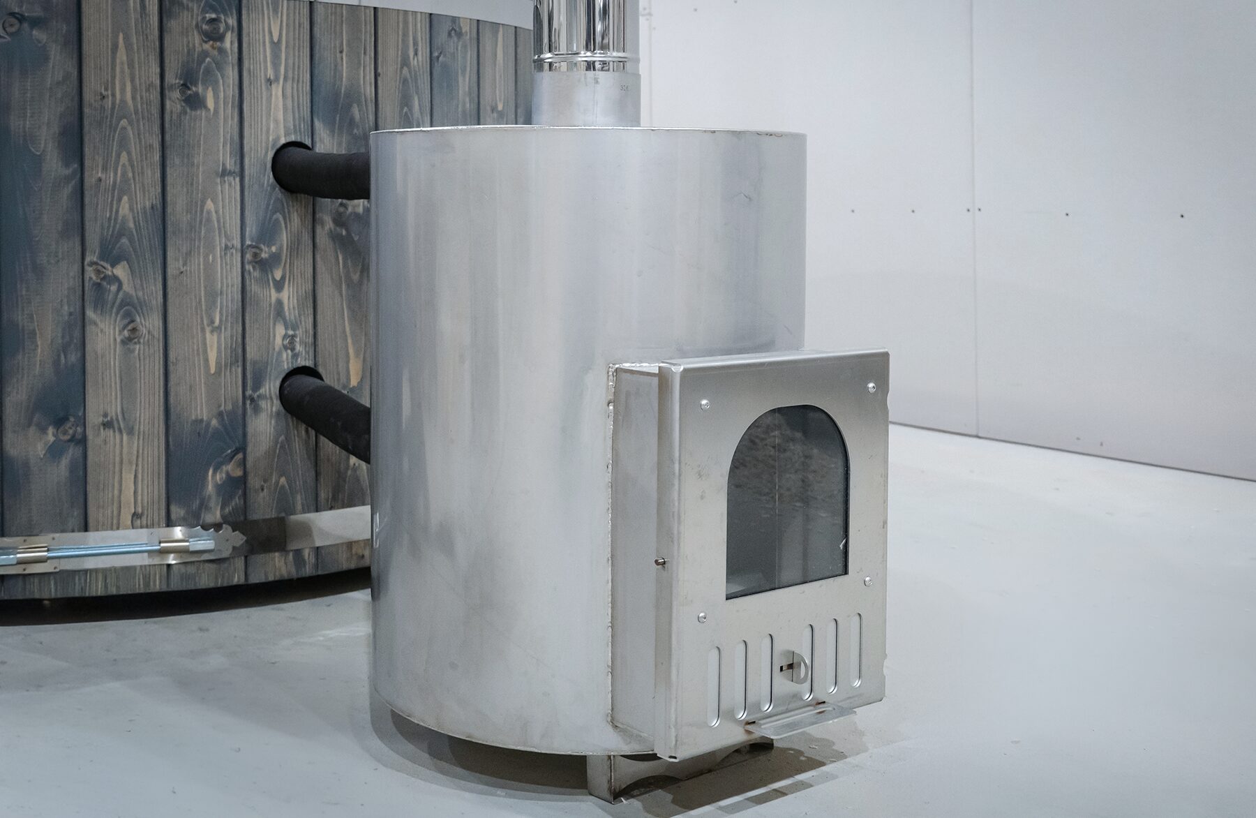 Close-up view of an external metal stove of the Makoya Spa hot tub DIANA, featuring the firebox door, connection pipes to the tub and a robust construction next to the wooden cladding